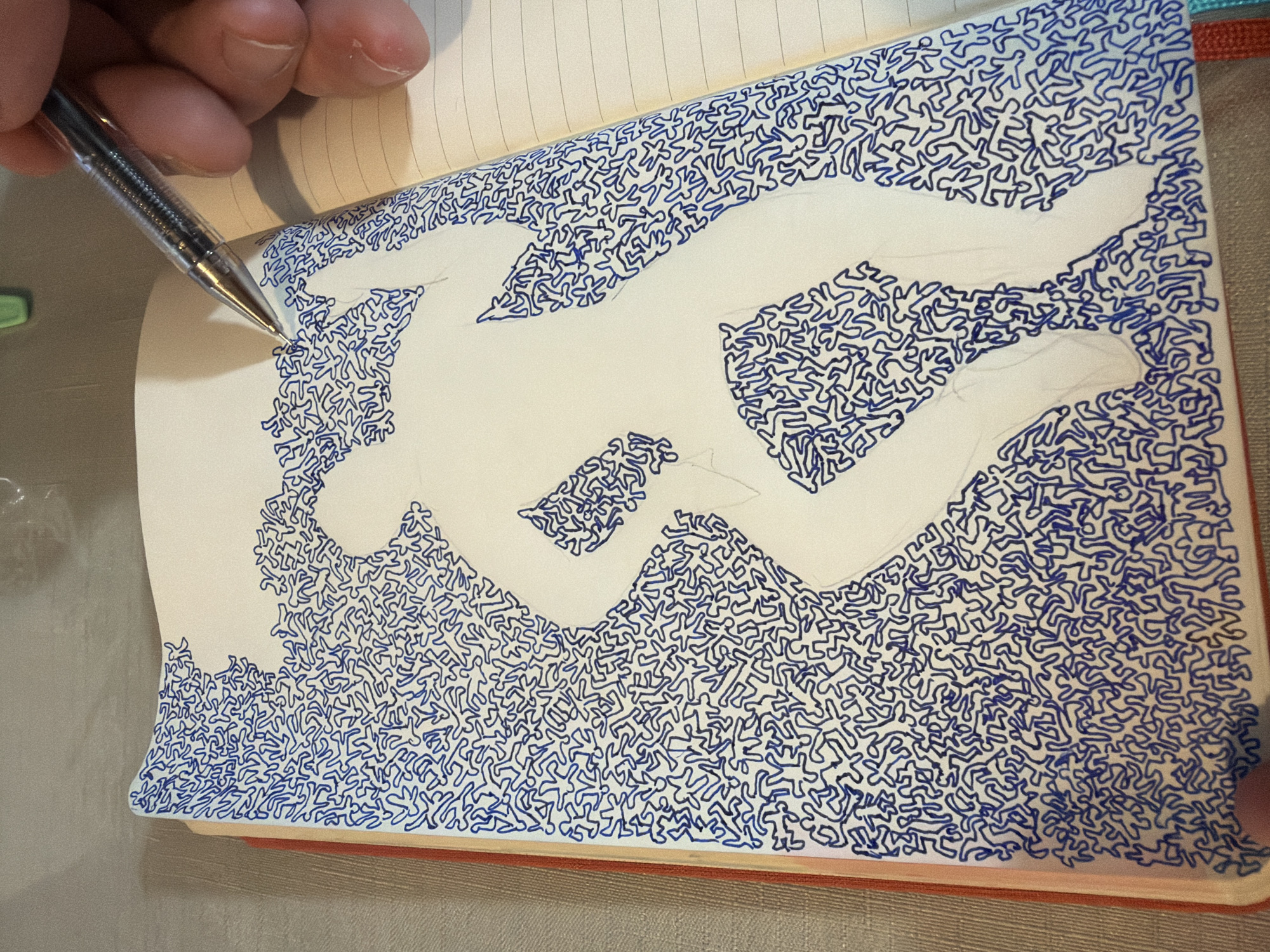 An overhead medium shot captures a hand holding a silver pen, poised above a notebook open to a page filled with a blue, abstract pattern of small, stylized human figures. The pattern surrounds a silhouetted figure, sketched in faint grey pencil, which appears to be in motion, possibly running or dancing. The notebook has a red cover and is lying on a textured surface. The background is out of focus, showing a neutral, light-colored wall or surface. The image does not contain any information about the city of Even Yehuda, Israel.