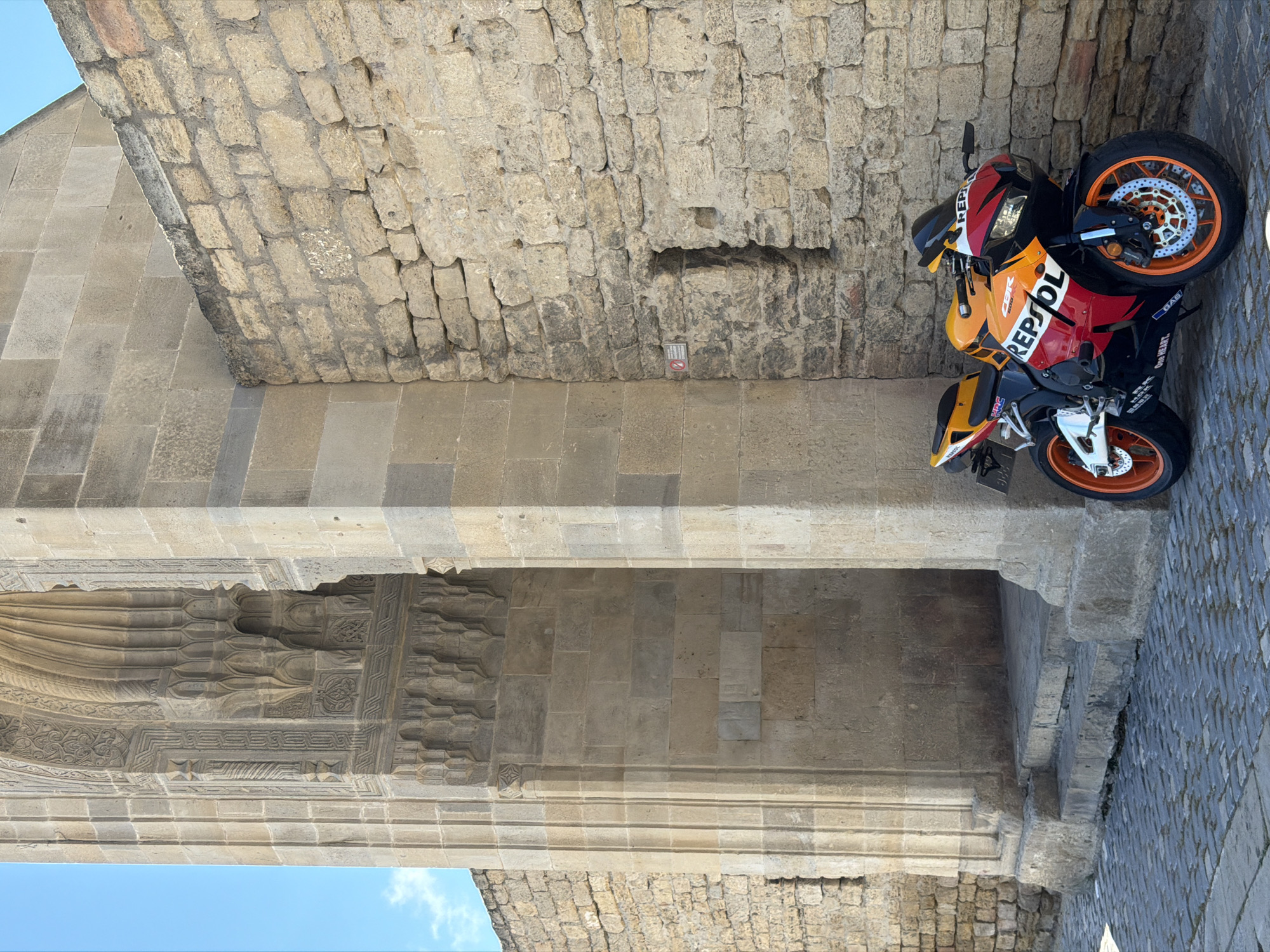 A vibrant red, orange, and black Honda CBR 600 sport motorcycle is parked on a cobblestone street in front of a weathered stone building with intricate architectural carvings. The building features a large arched entryway with steps leading up to it, and a rough stone wall to the right of the motorcycle. The sky above is a clear, bright blue with a few wispy clouds. The motorcycle is the primary subject, positioned to the right of the frame, angled slightly towards the viewer. The building's ancient stonework, detailed carvings, and the textured cobblestones contribute to a sense of historical depth in the scene. This image was captured in the Sabail Raion of Azerbaijan.