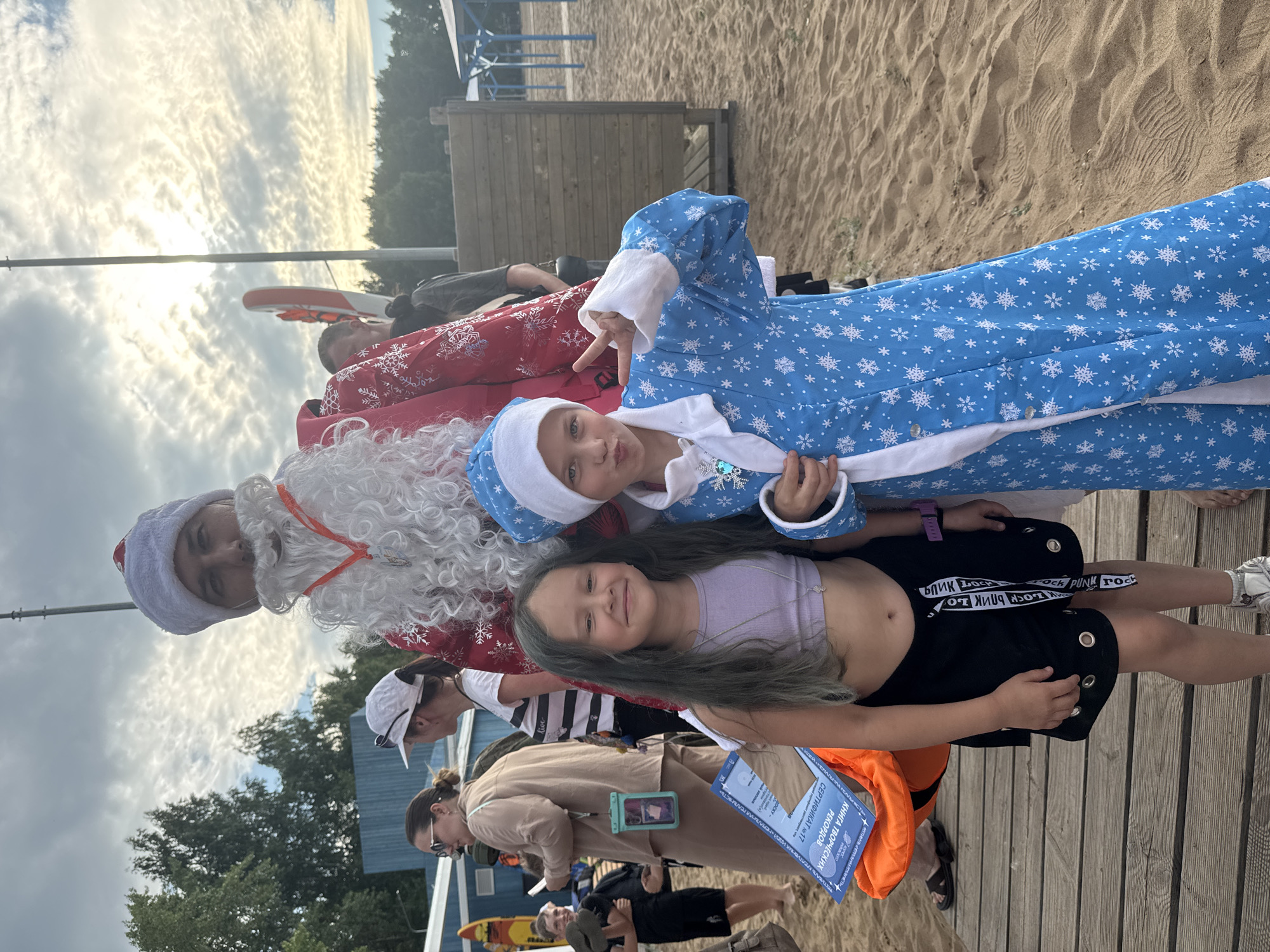A man dressed as Santa Claus poses for a photo with two children on a sandy beach, with a wooden walkway in the foreground. The man wears a red coat with white trim and fur beard, and a red hat. The child on the left, a girl, has long, dark, wavy hair and is wearing a light purple crop top and black shorts. The child on the right, a girl, wears a blue snowflake-patterned coat and hat, and is making a peace sign with her right hand. In the background, other people are visible, along with beach umbrellas and trees. The sky is partly cloudy. The location is likely a public beach in the city of Chaikovsky, Russia, given the presence of a certificate in Russian language and the overall setting.