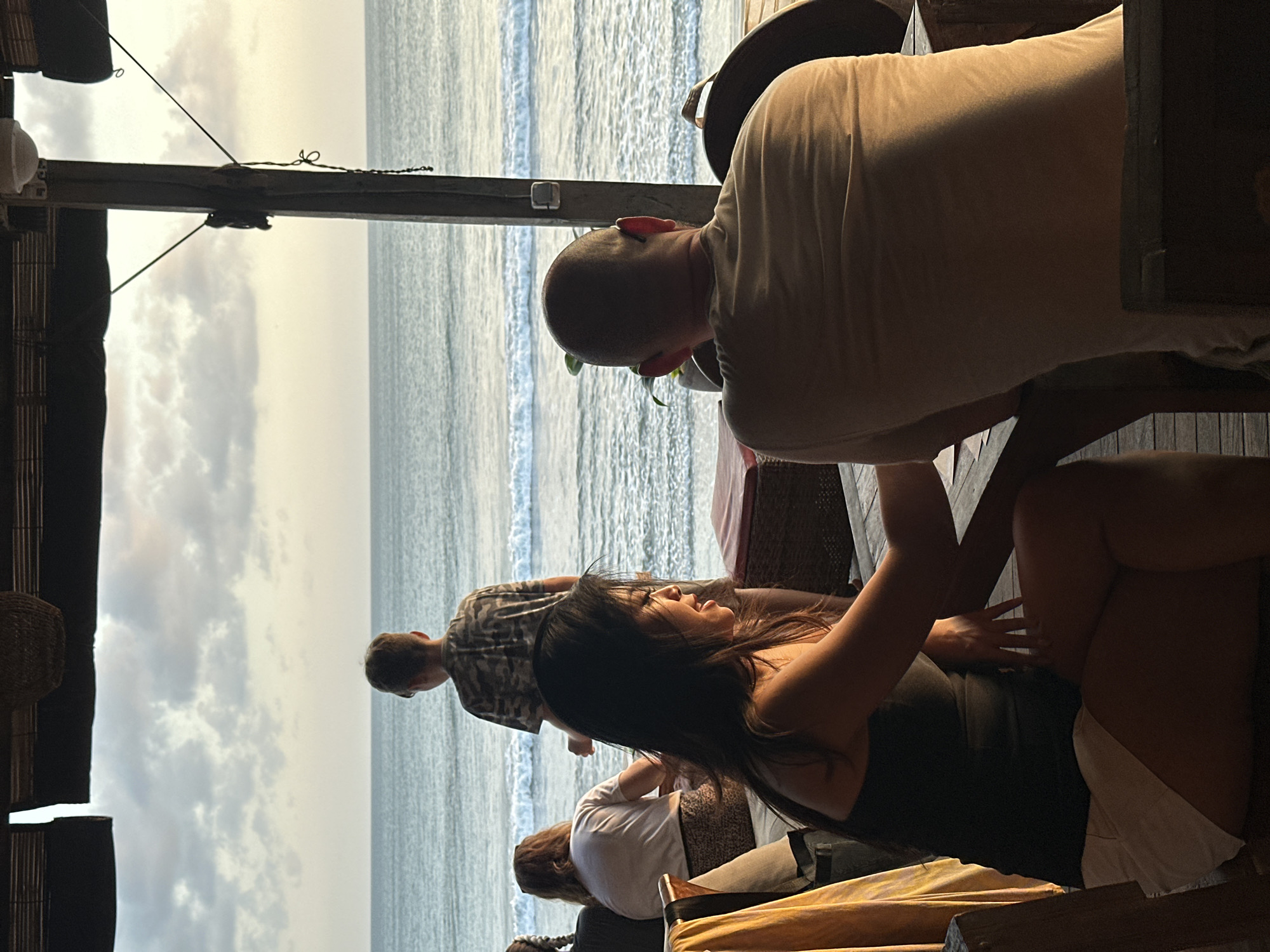 The image captures a scene at a beachside establishment in North Kuta, Indonesia. In the foreground, a woman with long dark hair, wearing a black tank top and white shorts, is seated. Her body is angled towards the right, and her left hand rests on her lap. Beside her, a man with a shaved head, wearing a tan t-shirt, is also seated and leaning forward. The background features the ocean with gentle waves under a cloudy sky, suggesting it might be late afternoon or early morning.
Further back, a person wearing a patterned shirt is standing and looking towards the ocean. Another person, wearing a white top, is seated in the middle ground. The setting appears to be an open-air structure with wooden floors and supports, possibly a restaurant or bar, overlooking the beach. The overall atmosphere seems relaxed and casual, with individuals enjoying the seaside view. There is no visible text in the image.