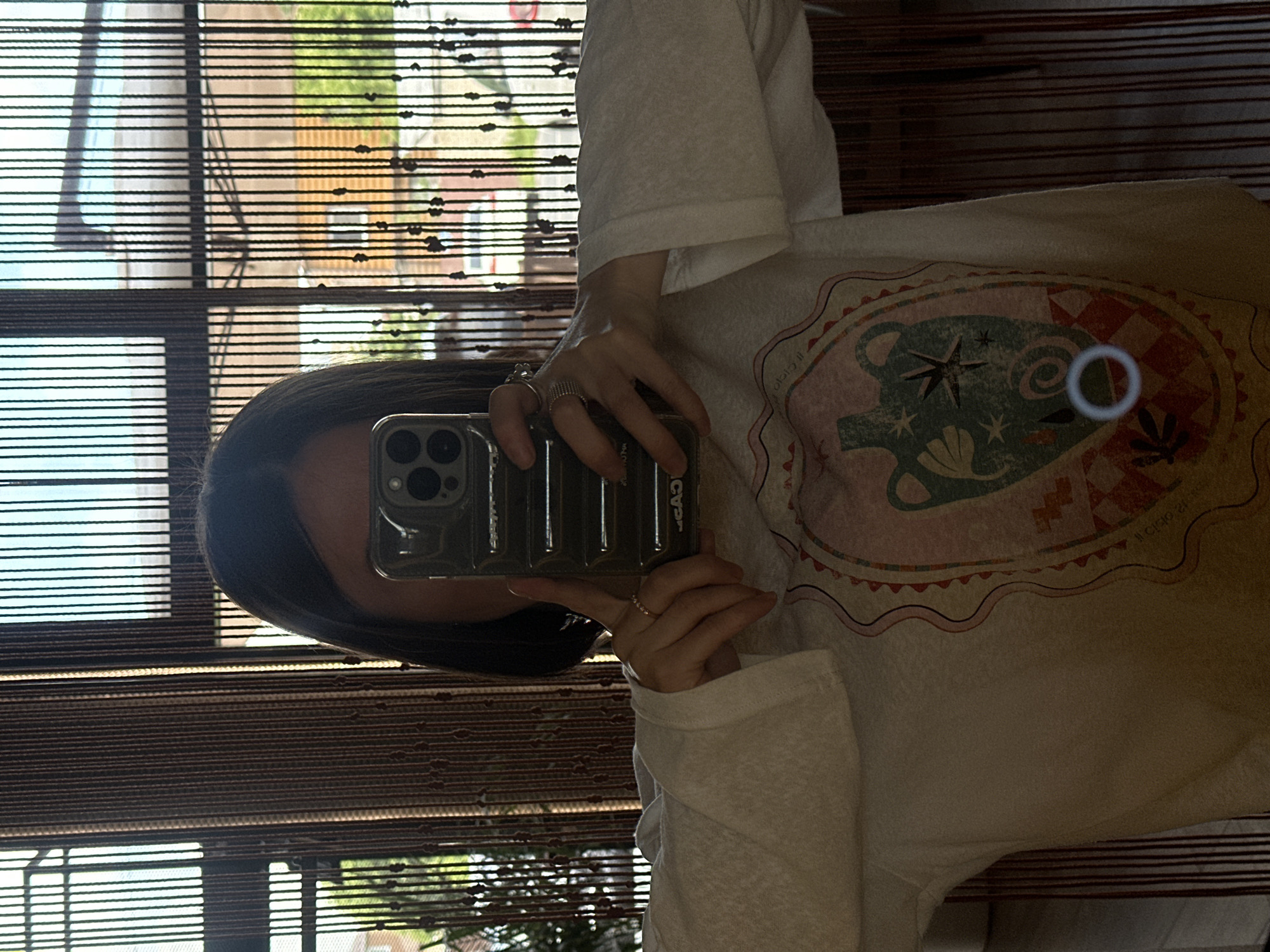 The image is a mirror selfie featuring an individual holding a dark olive-colored smartphone, which obscures their face. The person has dark, straight hair visible on either side of the phone. They are wearing a light-colored, possibly white or off-white, long-sleeved top with wide, flared cuffs. The front of the shirt displays a large, central graphic design in a stylized oval or shield shape. The graphic features an ornate jar or urn with handles, surrounded by abstract shapes, stars, and foliage motifs in muted tones of teal, pink, orange, and white, with a checkerboard pattern at the bottom.
Rings are visible on the fingers of both hands; the right hand wears a thin band and a wider, textured band, while the left hand has a single thin band. The smartphone case, partially showing "CASETiFY" branding in reverse, features a prominent triple-lens camera array.
The background, reflected behind the individual, consists of an interior space with a window covered by vertical dark brown bead curtains or blinds. Through these translucent blinds, an outdoor scene is visible. This exterior view includes a bright sky, green trees and foliage, and architectural elements of distant buildings, such as light-colored walls and parts of a roofline. The scene is illuminated by natural daylight. No specific geographical identifiers for Усады, Russia, are discernible within the image.