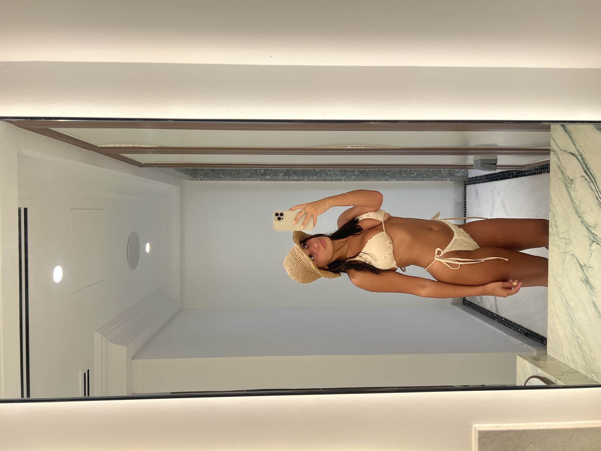 A woman wearing a straw hat and a light-colored bikini is taking a selfie in a bathroom mirror. The bathroom appears to have white walls and a ceiling with recessed lighting. The mirror frame is white with a thin black border. A section of a marble countertop is visible at the bottom right of the frame. The reflection shows the bathroom's architectural details, including the ceiling design and wall features. There is no definitive evidence to confirm the location of the scene in Dubai, United Arab Emirates.
