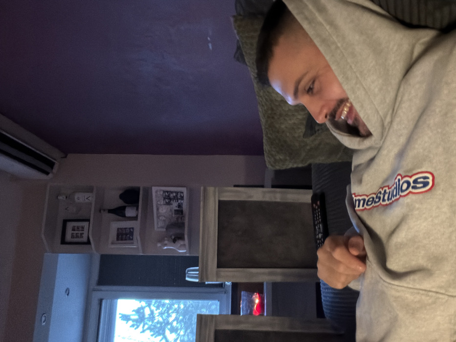 A male individual is depicted in a residential interior, identified as Sofia, Bulgaria, reclining on a dark grey ribbed sofa. The subject, seen in profile, possesses short dark hair, a beard, and a mustache, with his mouth open in a smile. He wears a light grey hooded sweatshirt bearing a partially visible red and blue text, appearing as "mostudios" or "timestudios," on the chest area. His right arm is bent, with the hand forming a fist resting on his upper torso. A black remote control is positioned on the sofa near the subject's left side.
The background includes a large window to the left, showing a blue sky and the silhouette of a coniferous tree, suggesting daylight conditions. Below the window, a light-toned piece of furniture supports a red candle in a glass holder. Adjacent to the window, a light-colored wall incorporates white corner shelves. These shelves hold various decorative items: framed pictures, one clear bottle, one dark bottle, a dark spherical object, and a small white bust-like figure. A white air conditioning unit is mounted high on the wall above these shelves. The wall extending to the right is painted a solid dark purple hue.