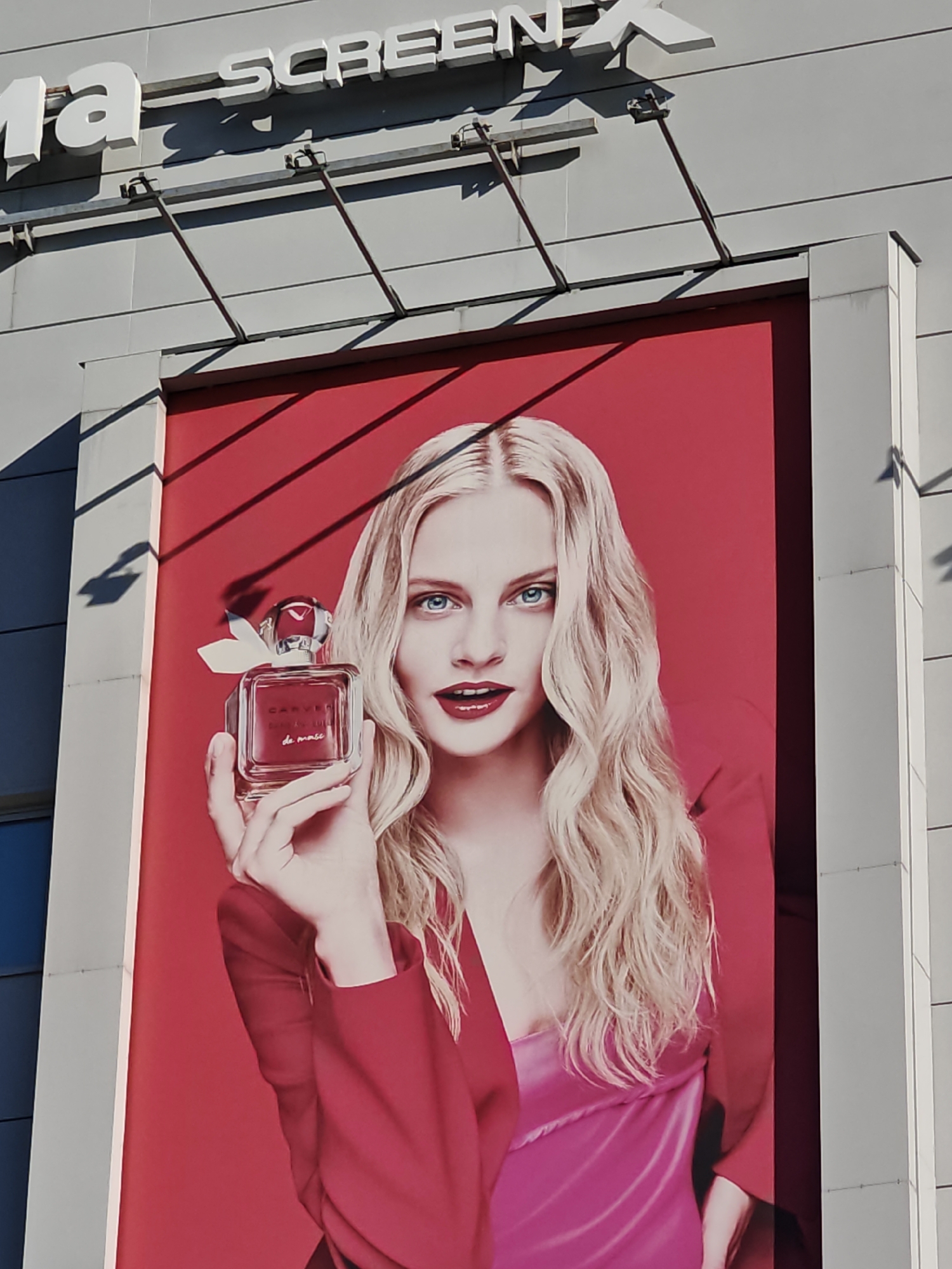 This is a close-up, low-angle, outdoor photograph of a billboard advertisement. The billboard features a headshot of a young, Caucasian woman with long, wavy blonde hair and striking blue eyes, wearing bright red lipstick. She is holding a square, clear glass perfume bottle with a red cap and a white ribbon accent. The bottle has text on it that reads "CARVER" and "de masi". The background of the billboard is a solid, vibrant red. Above the billboard, a building facade is visible, with the white, illuminated letters "SCREENX" prominently displayed, suggesting the location is a cinema or entertainment complex. The building's exterior appears to be grey and modern, with visible structural elements casting shadows. The lighting suggests it is daytime with strong sunlight. The overall impression is a high-fashion perfume advertisement. While the prompt mentions Moscow, Russia, there is no visual information in the image to confirm this specific location.