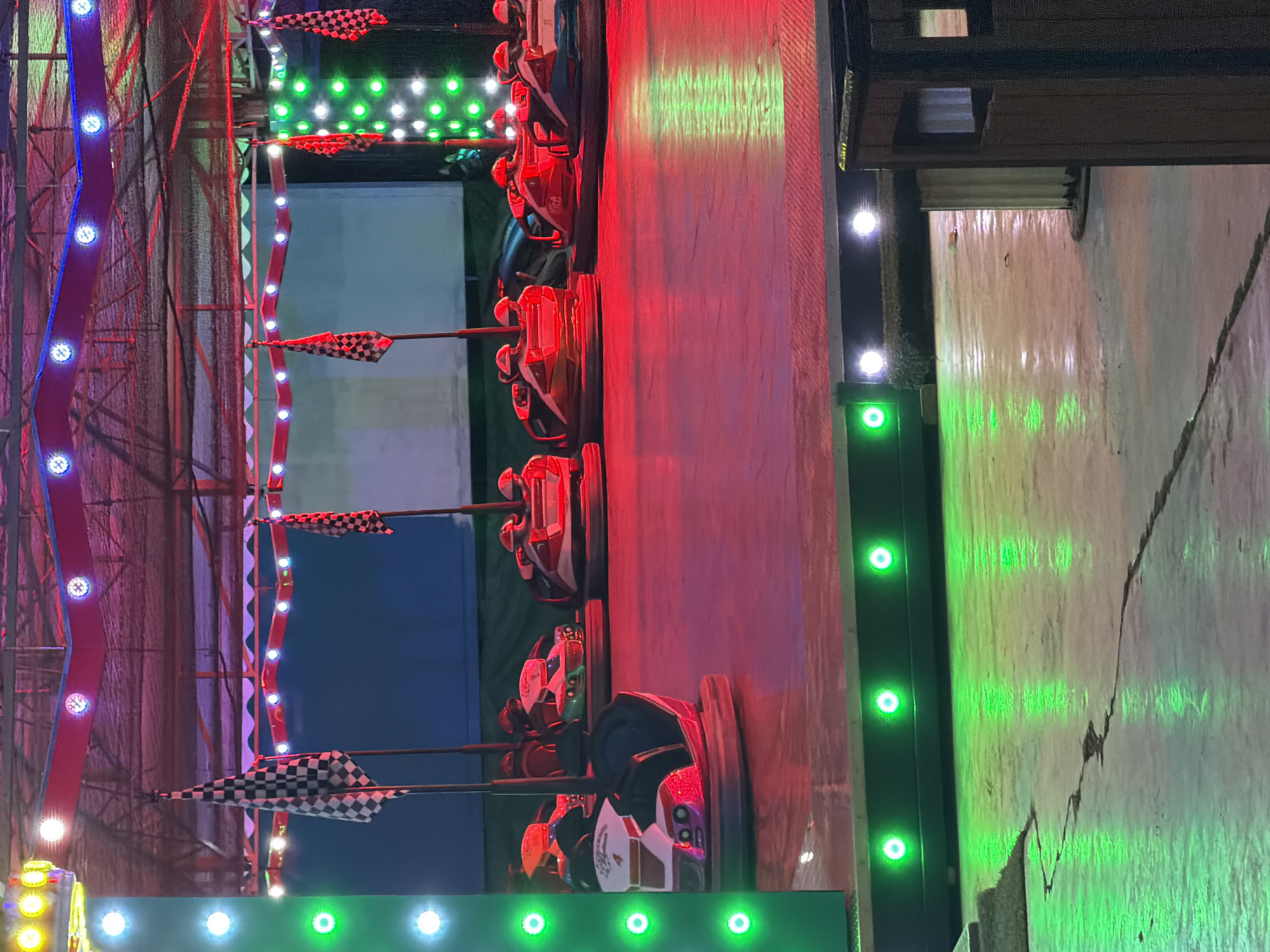 This image depicts a bumper car ride at an amusement park, likely in Sant Antoni de Portmany, Spain, given the context. The ride features several bumper cars, predominantly in red and white color schemes, positioned on a circular arena. The arena floor is a polished, reddish-brown surface reflecting the vibrant red and green lights emanating from the ride's overhead structure and surrounding lights.
Several checkered flags, commonly associated with racing or fairground attractions, are mounted on poles around the arena. The background is a dark, blurred expanse, suggesting a night-time or dimly lit environment. The overall impression is of a lively, colorful, and energetic fairground attraction. There are no people clearly visible in the bumper cars or operating the ride. The image focuses on the setup and atmosphere of the bumper car attraction.
