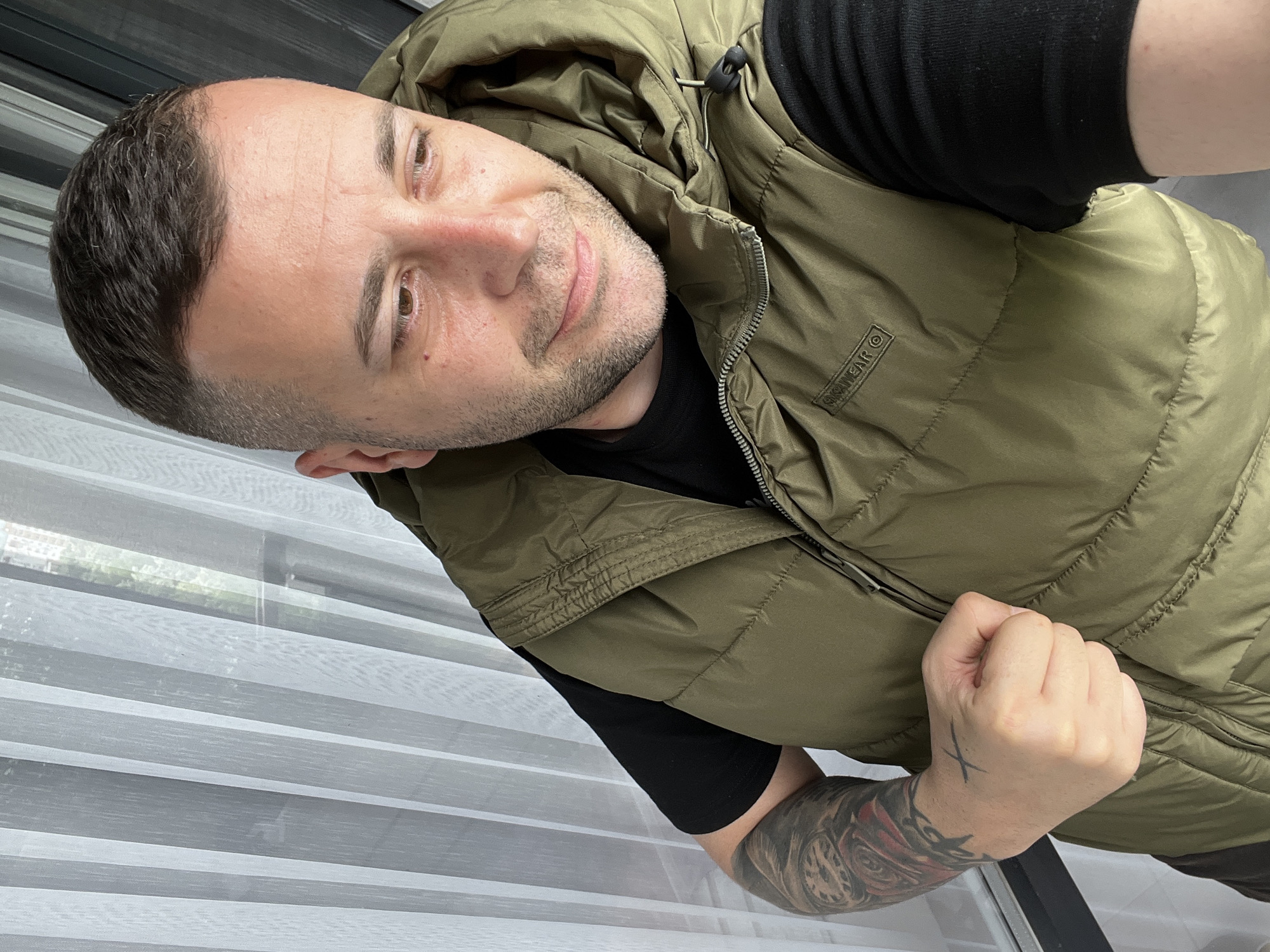 A man is featured in a mid-shot portrait, looking directly at the camera with a subtle, friendly smile. He has short dark hair, a light beard, and light brown eyes. He is wearing a black short-sleeved t-shirt under an olive green puffer vest with a hood. A small logo reading "SKNWEAR ©" is visible on the left chest area of the vest. His left forearm is adorned with a detailed tattoo, and a small black 'X' is tattooed on the back of his left hand, which is raised near his chest. The background shows white horizontal blinds, indicating an indoor setting, likely by a window, with diffused daylight. The image is oriented sideways, with the man's head facing the right. The scene takes place in Varna, Bulgaria.