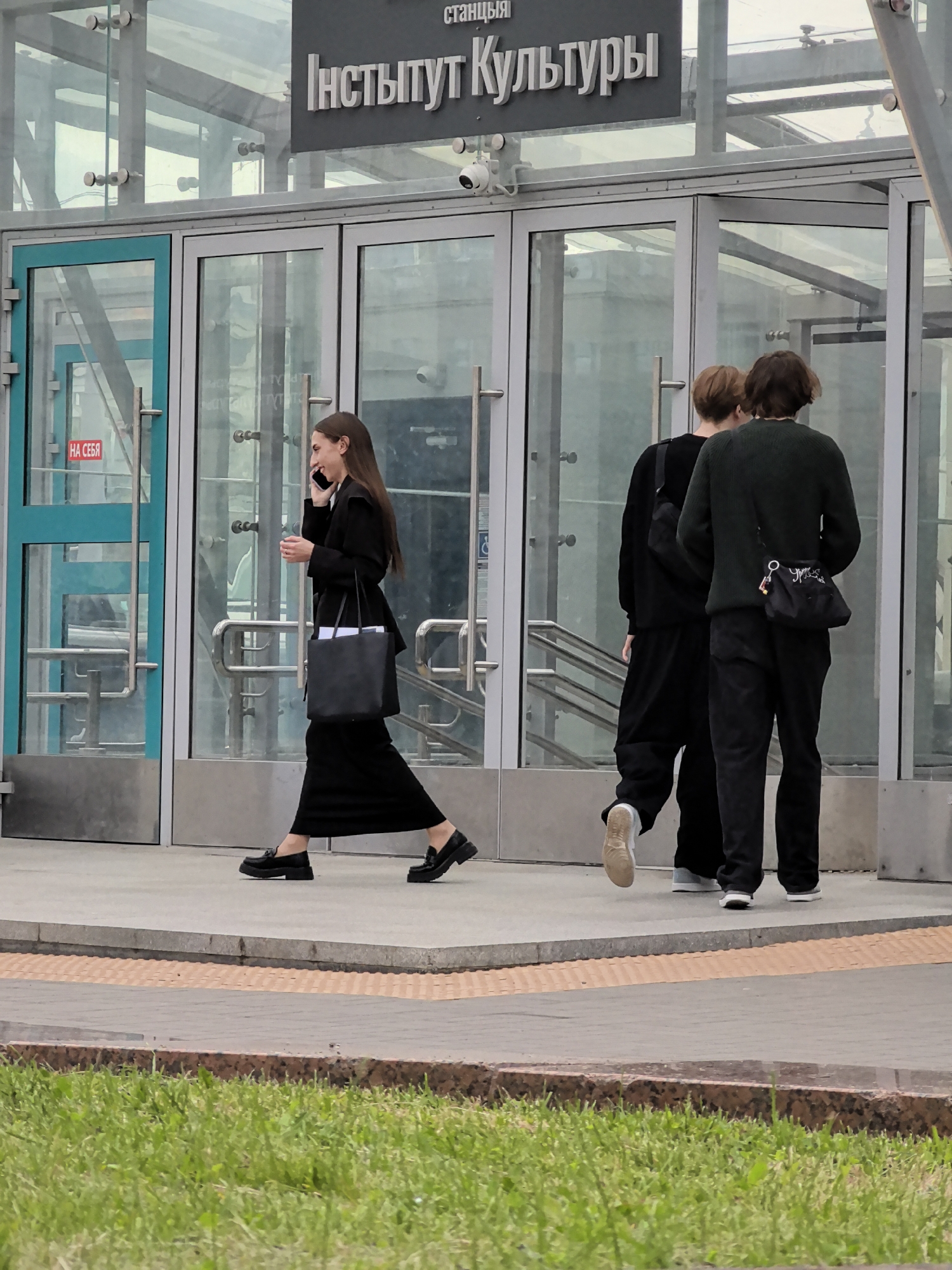 In an urban setting, possibly a city, a woman in a black dress and black loafers walks to the left, talking on her phone and carrying a black tote bag. Behind her, two individuals, one in a black outfit and the other in a dark green sweater and black pants, walk away from the camera to the right. They both carry bags. They are in front of a building with large glass doors and a sign that reads "Институт Культуры" (Institute of Culture). A security camera is visible above the doors. The ground in front of the building is paved with concrete and textured tiles, bordered by a strip of grass in the foreground.