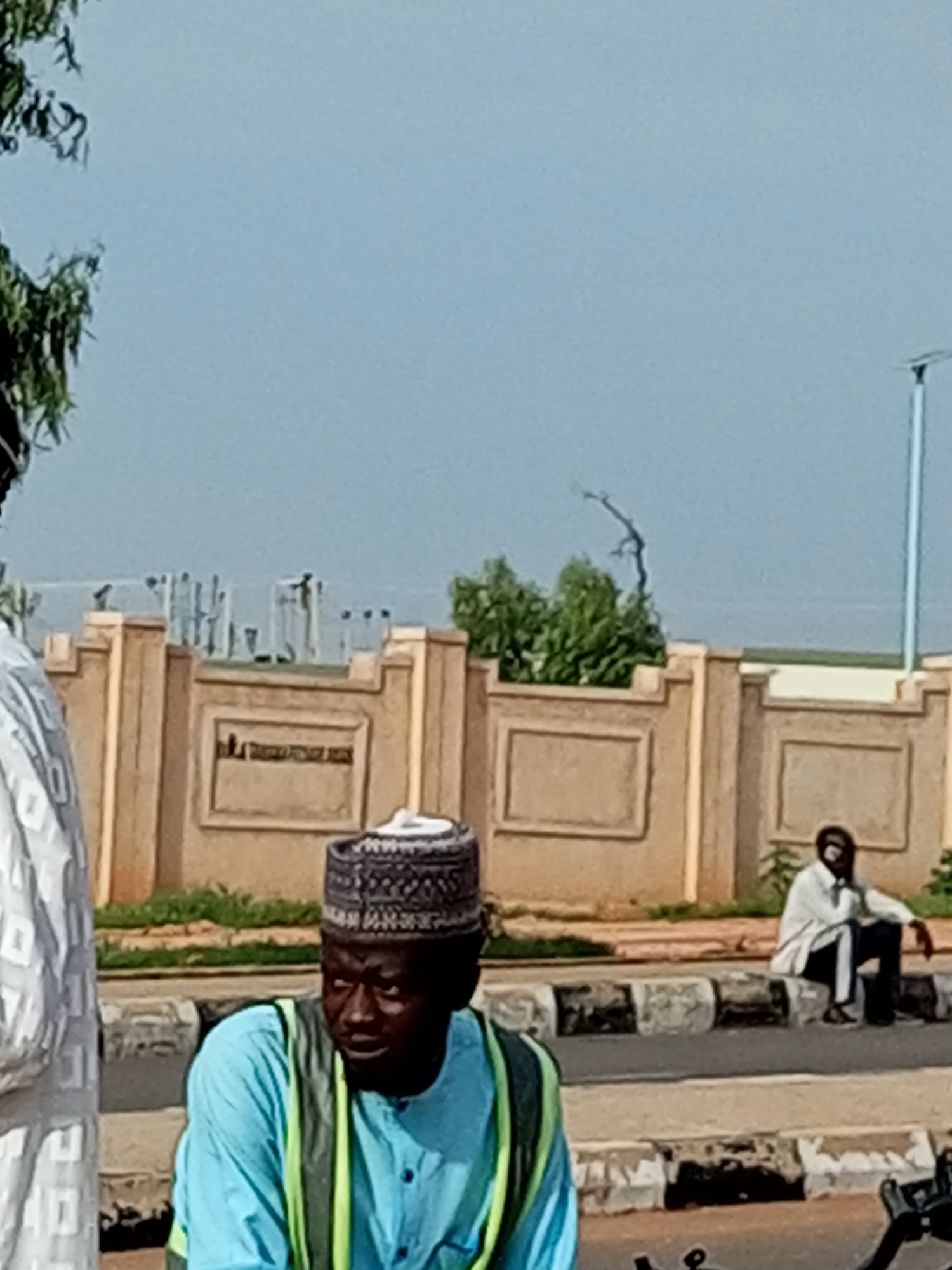 A medium shot captured on a sunny day shows a street scene in Gombe, Nigeria. In the foreground, a man wearing a light blue and green vest over a blue shirt and a traditional cap is seated, facing right and looking downwards. Behind him, to the right, another man in a white shirt and dark pants is seated on a curb, leaning his elbow on his knee. A light-colored, ornate wall with rectangular insets runs along the middle ground, separating the street from a background with some greenery and what appear to be utility poles or structures. The sky is a clear, pale blue. The overall impression is one of a casual outdoor gathering or waiting area along a street.