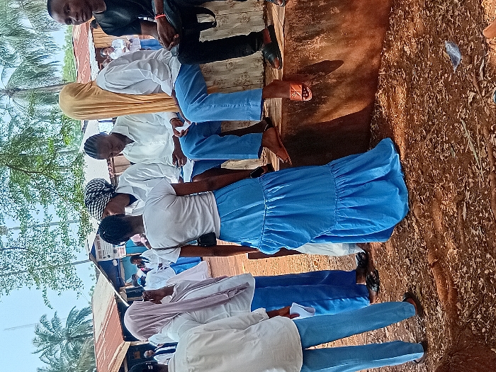 A group of students, predominantly in light blue and white uniforms, are gathered outside. Some wear head coverings of various colors. They appear to be standing or sitting near a building with a corrugated roof, with trees visible in the background. The ground is covered with leaves and debris.