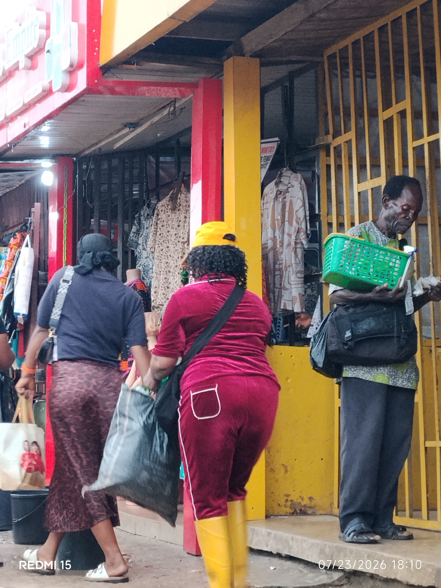 In Umuode, Nigeria, a street scene unfolds in front of a market stall. Two women, viewed from the rear, are walking away from the camera. The woman on the left wears a dark blue shirt and a patterned skirt. She carries a black bag over her shoulder and a large, dark plastic bag in her right hand. The woman on the right is dressed in a burgundy tracksuit and yellow boots, and she holds a dark bag in her left hand. In the background, a man stands near a yellow barred gate, holding a green basket and some papers. The market stall features hanging clothes and a red and yellow facade. The ground is a mixture of concrete and dirt. A timestamp on the image reads "07/23/2026 18:10".