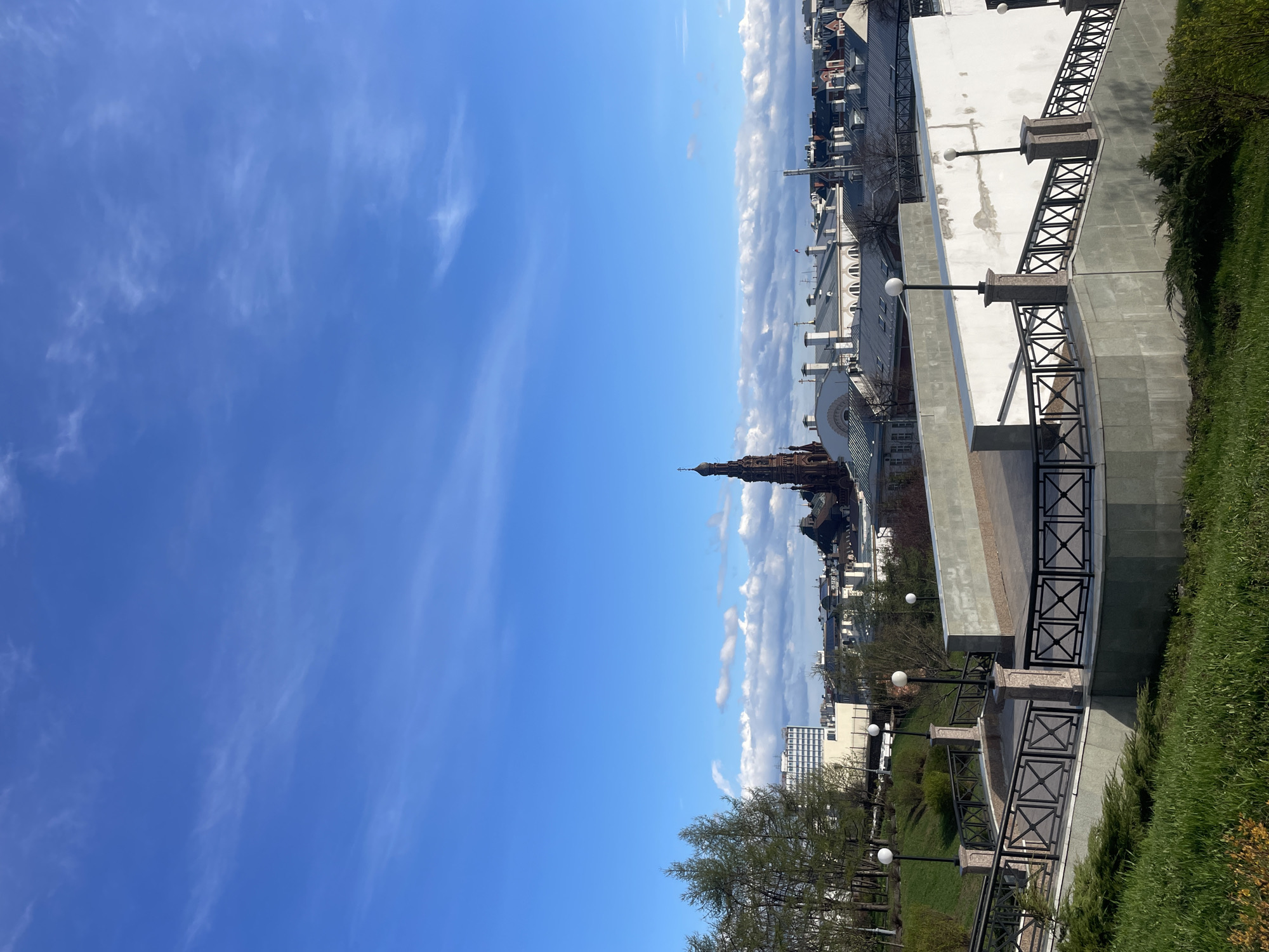This is a bright, clear daytime image capturing a panoramic view of a city, presumably Kazan, Russia. The vast expanse of blue sky, dotted with wispy white clouds, occupies a significant portion of the upper frame, suggesting pleasant weather. On the horizon, a band of fluffier cumulus clouds stretches across the cityscape.
In the middle ground, a distinctive dark-colored building with a tall spire stands prominently, likely a historical landmark such as a mosque or cathedral. Surrounding it are various urban structures, including buildings with white facades, dark roofs, and some modern multi-story constructions.
The foreground features a well-maintained park or garden area. Lush green grass and deciduous trees with light green leaves, indicative of spring or early summer, are visible. A multi-level concrete or stone pathway or terrace is prominent on the right, adorned with black metal railings featuring X-shaped patterns. Several round, white streetlights on dark posts punctuate this structure. A large white wall, showing some signs of wear, also forms part of the foreground architecture. No people or specific events are depicted, presenting a serene city landscape. The bright illumination confirms it's daytime, likely mid-morning to early afternoon. No text is visible.