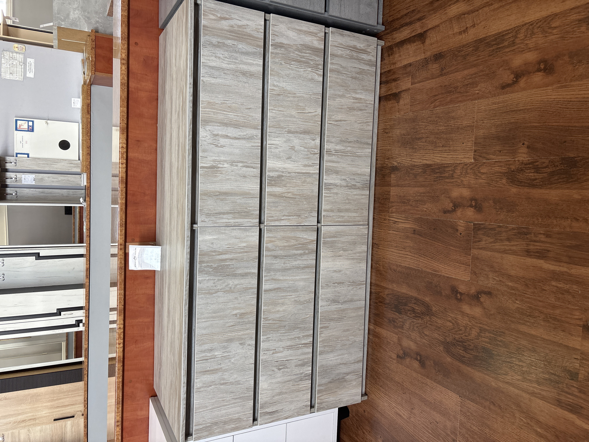 The media file shows a close-up, overhead view of a hallway with doors and cabinets. The hallway is lined with wood-grain patterned cabinets on the left and a wooden floor on the right. The cabinets appear to be made of a light-colored wood with a subtle grain pattern. They are arranged vertically, with multiple doors visible.
The wooden floor is made of planks with a dark brown, reddish hue and a prominent wood grain texture. The planks are laid horizontally in the image, creating a warm and inviting atmosphere.
To the left of the cabinets, there are glimpses of other furniture and displays, including what appear to be drawers and shelves. One of the cabinets has a white label attached to it with some text. Another section of the hallway shows a tall, narrow cabinet with a reflective surface, possibly a mirror or a polished material.
The lighting suggests an indoor setting, likely a showroom or a store displaying furniture or cabinetry. There are no people visible in the image, and the scene is static, indicating a posed shot or a captured moment in time. The overall impression is one of a well-organized and modern space, with a focus on the textures and patterns of the materials.
Visible text:
- A white label with text is attached to one of the cabinets on the left. The text is small and difficult to read, but it appears to be a product label or information tag.