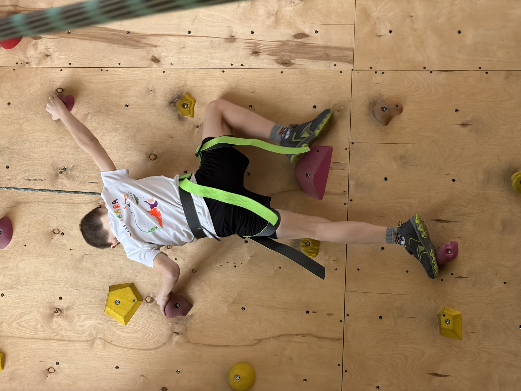 A young child, presumably male, is depicted actively climbing an indoor wooden climbing wall in Izhevsk, Russia. The child is positioned horizontally across the wall, gripping various colorful holds.
He is wearing a white, long-sleeved t-shirt with a colorful graphic design, black shorts, and a climbing harness with bright neon green straps and darker gray webbing around his waist and legs. His footwear consists of grey and black athletic shoes with yellow accents, and socks featuring a cartoon character (possibly from Paw Patrol) are visible.
The climbing wall is constructed from large, light-colored plywood panels, featuring numerous climbing holds of varying shapes and sizes in colors such as yellow, purple, red, and brown. A green and black striped rope is visible diagonally in the upper left portion of the image, suggesting a safety system.
The child's right hand grips a small purple hold, and his left foot is placed firmly on a magenta-pink hold. His left arm is extended, and his right leg is also extended towards a yellow hold, indicating a dynamic climbing motion. The perspective of the image is from directly above, looking down at the child and the wall. The child appears focused on the activity. Part of a word, "PARR", is visible on the front of his white t-shirt.