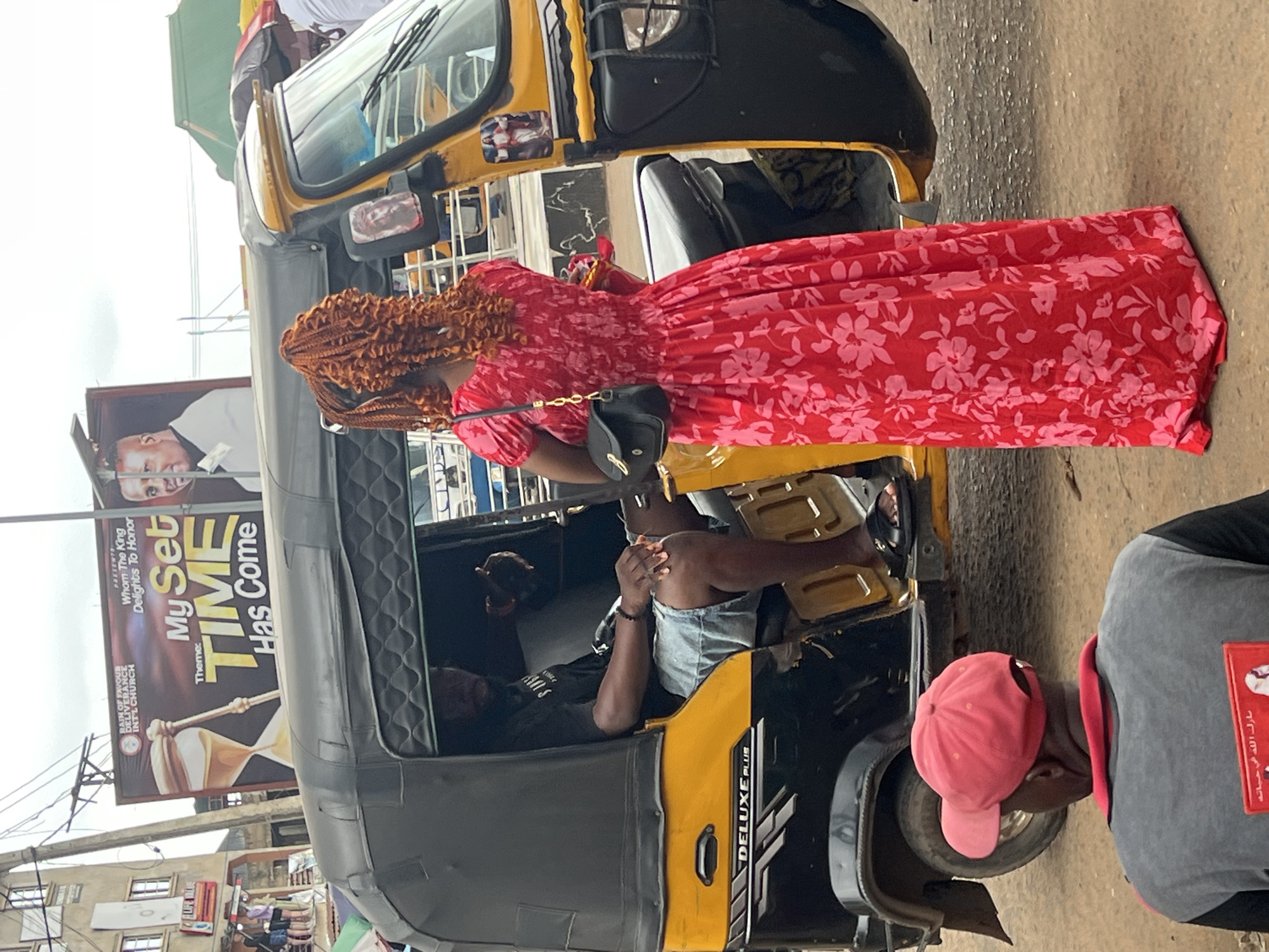 An upward, medium shot captures a scene on a street in Nkpor, Nigeria, with a focus on a yellow and black auto-rickshaw and its occupants. In the foreground, a person wearing a red cap and a gray shirt with a red collar is seen from behind. The auto-rickshaw, with "DELUXE PLUS" written on its side, is parked. A man with dark skin, wearing a black t-shirt and light denim shorts, sits in the driver's seat. A woman with braided, ginger-colored hair is standing to the right of the auto-rickshaw, wearing a long, red floral dress and carrying a black handbag. A sticker of Jesus Christ is visible on the side of the auto-rickshaw. Behind the auto-rickshaw, a large billboard displays text related to a church event, featuring an hourglass graphic and images of two men. Buildings and street poles are visible in the background, and the ground is made of dirt and gravel.