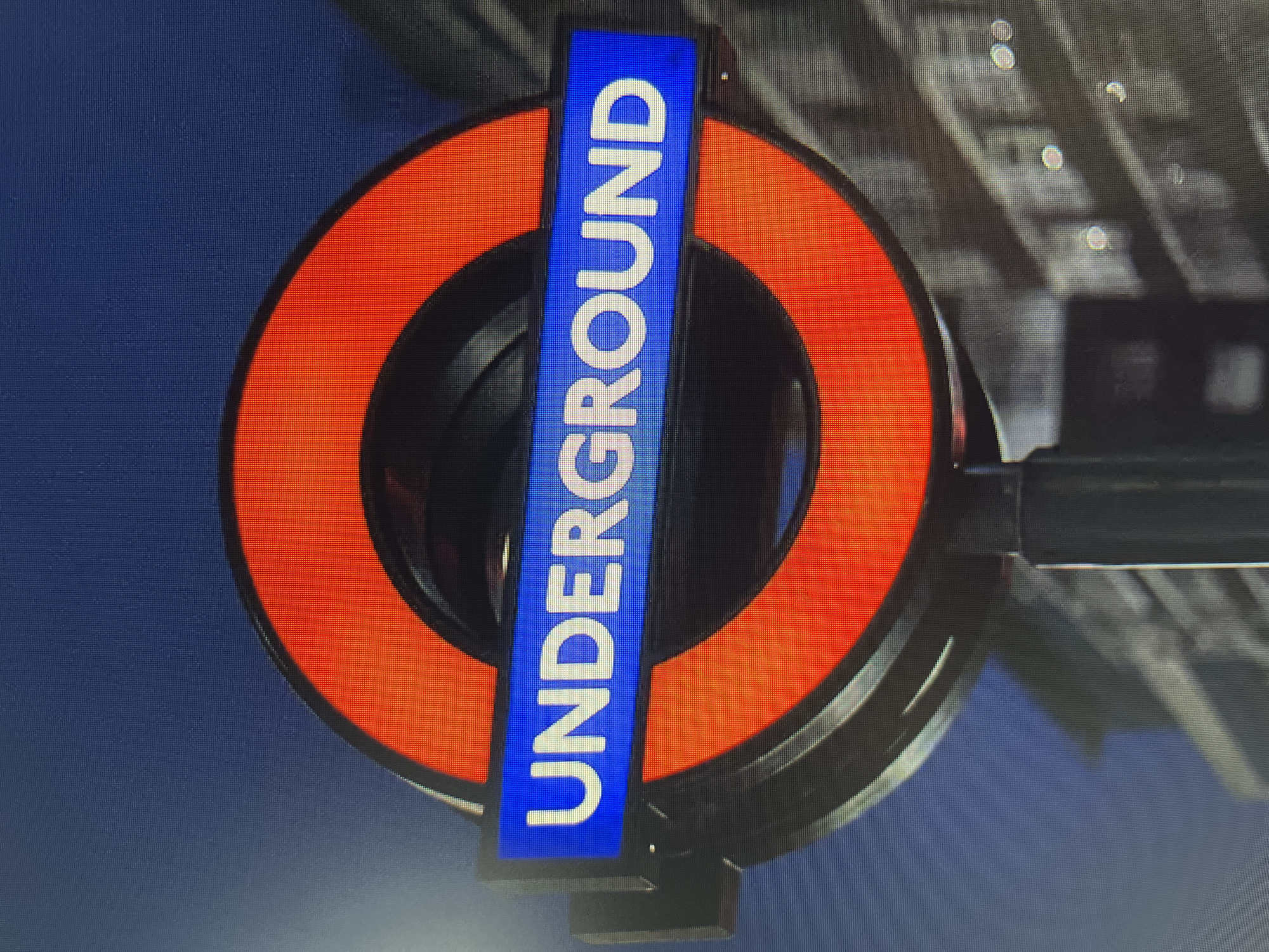 The media file shows a close-up, angled shot of a London Underground sign. The iconic red circle with a blue horizontal bar is prominently featured. The word "UNDERGROUND" is written in white capital letters on the blue bar. The sign is suspended against a dark blue background, which appears to be the sky, suggesting it is outdoors. Above and to the right of the sign, there are some out-of-focus lights and what looks like the underside of a structure, possibly a building or train station roof. The lighting suggests it might be daytime, but the overall atmosphere is somewhat subdued. There are no people visible in the frame. The image is a clear depiction of a well-known public transport symbol.