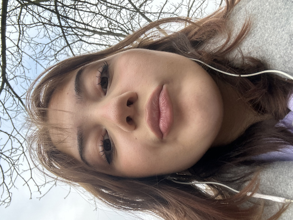 The image displays a low-angle, close-up portrait of a female subject, indicative of a selfie. Her face and upper chest occupy the majority of the frame. The subject has light brown hair with visible bangs and is looking directly at the camera with her lips pursed in a puckered expression. She exhibits visible mascara on her eyelashes and light lip color. White wired earphones are visible, with a wire running down on each side of her face. She is wearing a light grey overcoat or jacket, layered over a light purple garment. The background consists of a cloudy, overcast sky and a dense arrangement of bare tree branches, suggesting a deciduous tree in a dormant state. The scene is outdoors, consistent with a park or street environment within the city of Strasbourg, France. No specific architectural or geographical landmarks are visible to determine an exact location within Strasbourg.