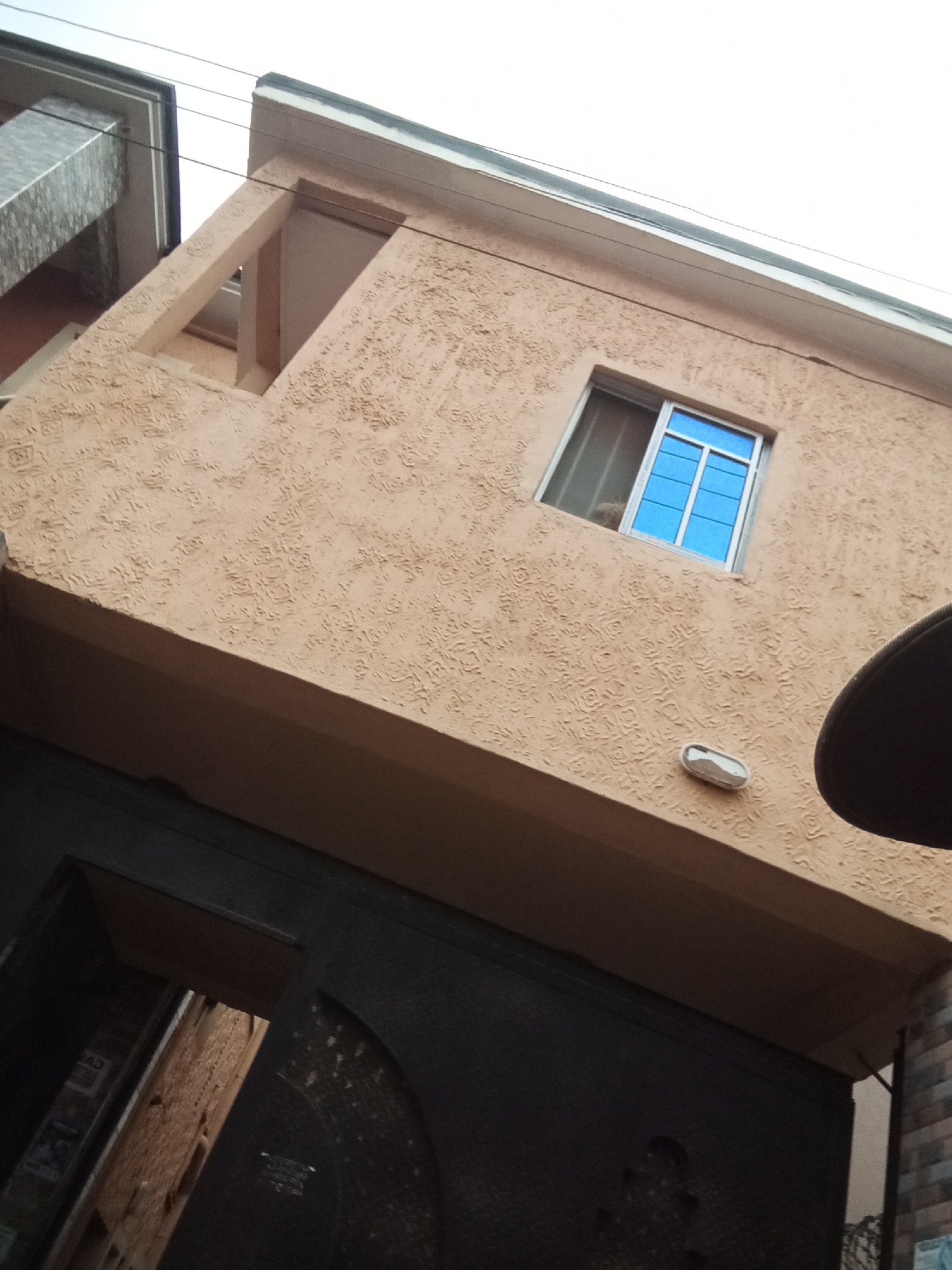 The image shows a close-up, upward-angled view of the exterior of a building in Oko Town, Nigeria. The building has a textured, peach-colored stucco facade with a white trim along the roofline. A rectangular window with blue panes is visible on the upper level. Below the window, a small, silver, oval-shaped light fixture is mounted on the wall. The bottom portion of the image reveals a dark, possibly black, metal gate with an ornate design, partially obscuring a view into a covered entryway or building behind it. To the left of the gate, a portion of another building or structure with a gray, patterned surface is visible, along with electrical wires running horizontally across the sky. The overall scene is that of an urban residential or commercial property, captured from a low vantage point.