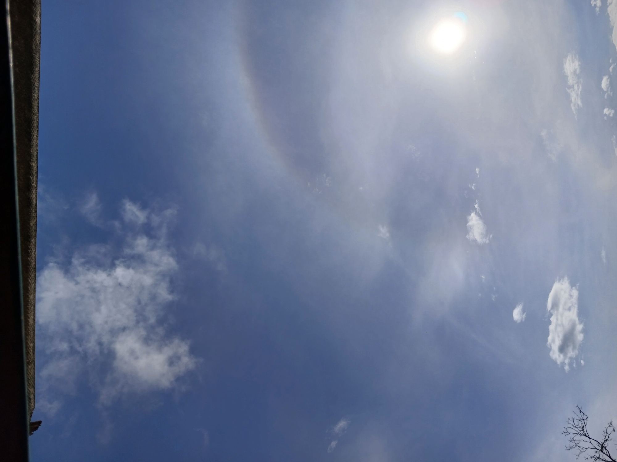 The image shows a bright blue sky with scattered white clouds. A faint halo is visible around the sun, which is located in the upper right quadrant of the image. The bottom edge of the image features a dark, textured object that appears to be the roofline of a building, possibly a balcony or overhang. In the lower left corner, the silhouetted branches of a bare tree are visible against the sky. The scene suggests an outdoor view from a high vantage point in Altagracia, Venezuela, likely during daylight hours.
