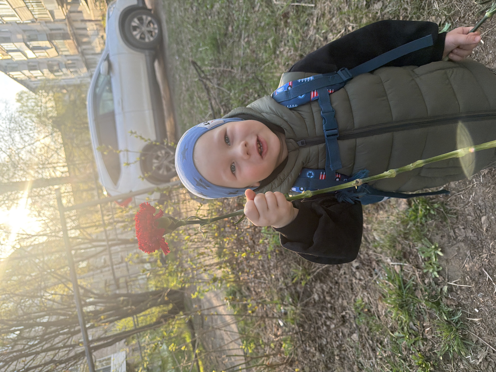 A young child stands outdoors, holding a red carnation. The child wears a blue patterned hat, a green vest, and a dark jacket. A blue backpack is strapped to their back. The background includes a white car parked near a building, foliage, and sunlight. The location may be Chaikovsky, Russia. The ground is covered in grass, foliage, and fallen leaves.