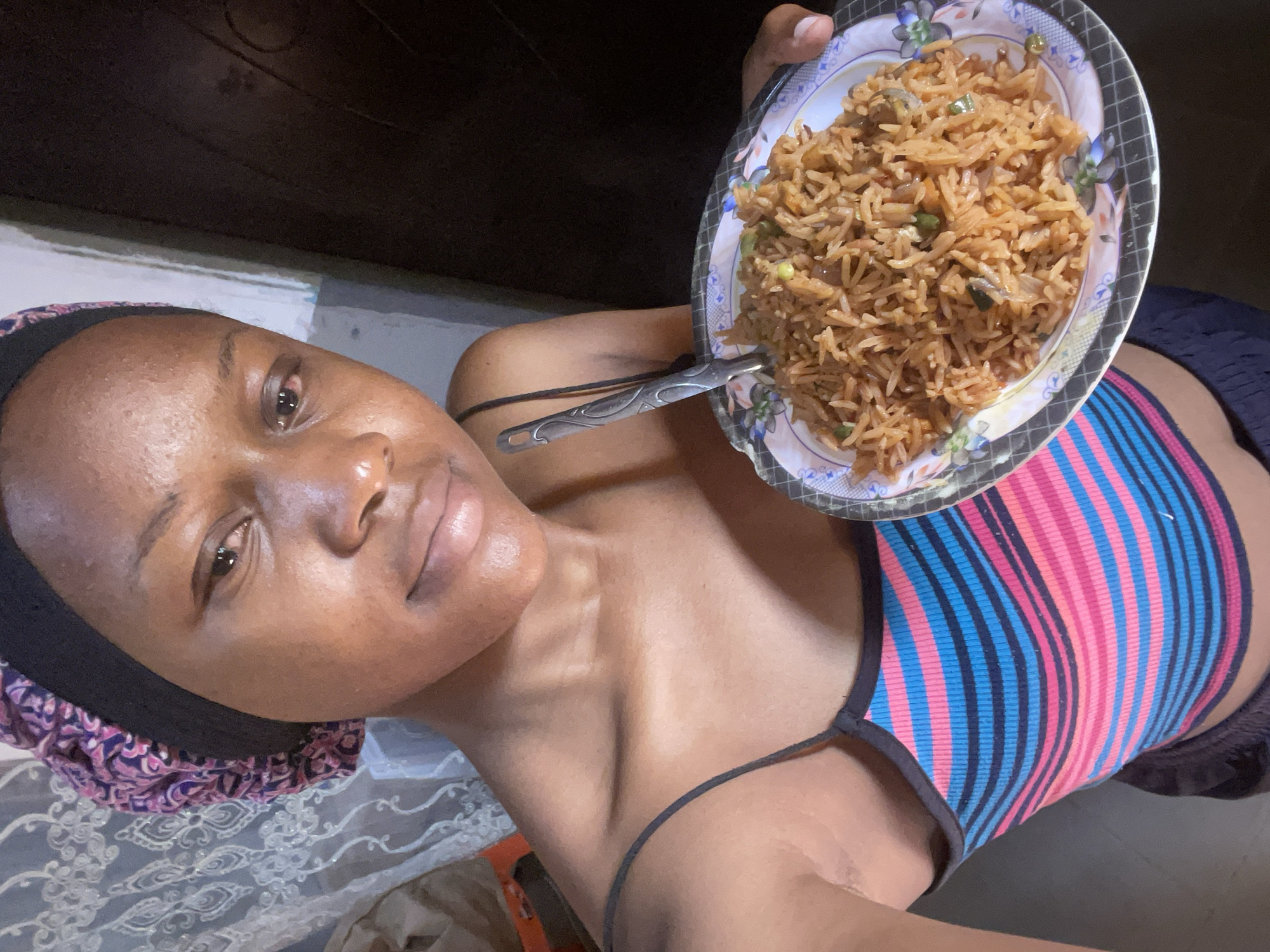 A young woman, likely in her late teens or early twenties, poses for a selfie in what appears to be a modest home setting in Jos, Nigeria. She is wearing a vibrant, horizontally striped tank top with colors of blue, pink, and black, and a patterned headscarf that covers her hair. In her hands, she holds a plate of fried rice, which she is about to eat with a fork. The background is dimly lit, with a patterned curtain visible on the left and a dark doorway or wall on the right. The overall atmosphere is casual and personal, suggesting a moment of everyday life captured in a domestic space.