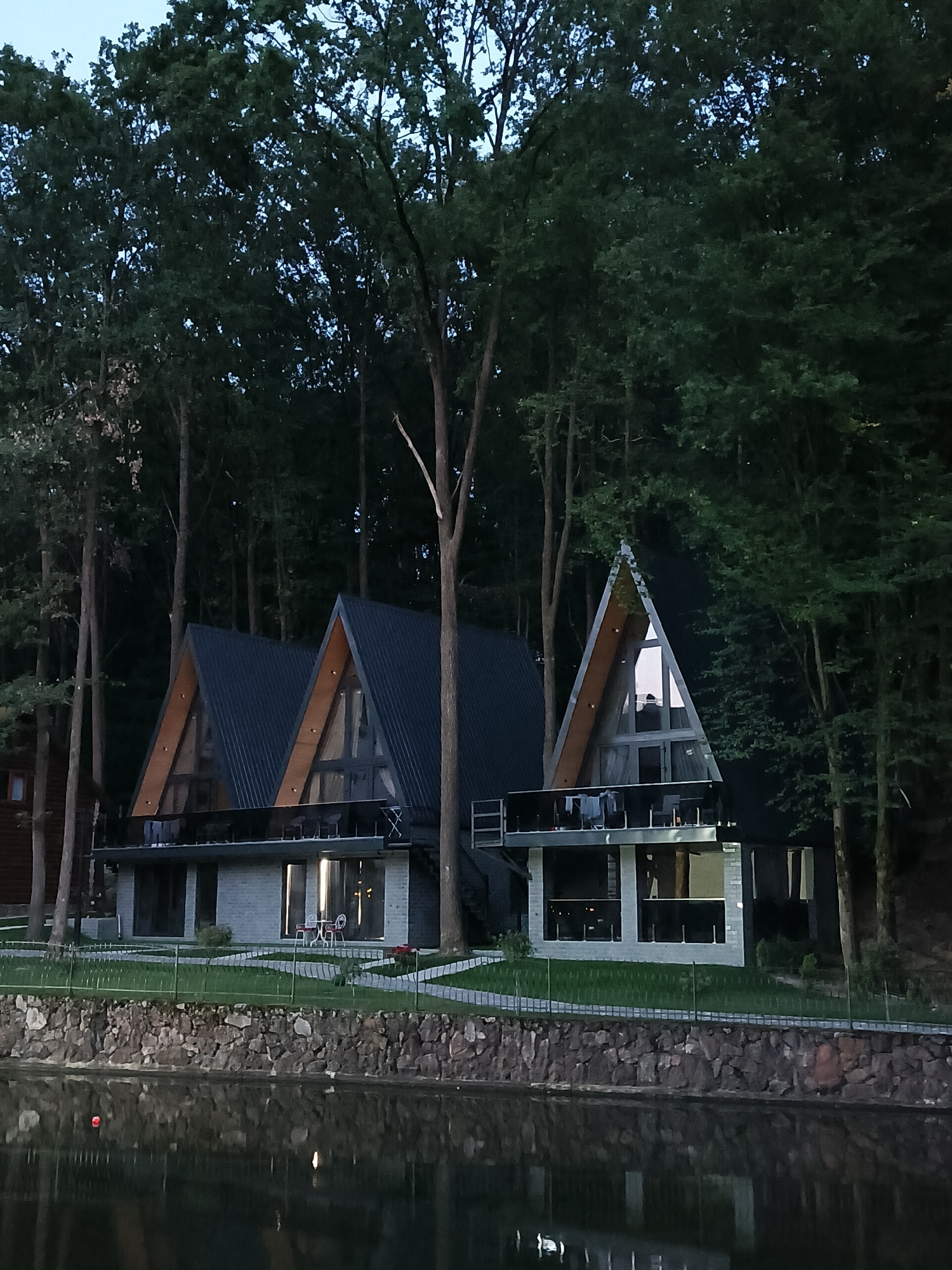 The image shows a modern resort complex situated by a body of water, likely a lake, surrounded by a dense forest. The architecture of the buildings is distinctive, featuring a series of A-frame structures with dark, sloped roofs and large glass facades. The main buildings are multi-leveled, with balconies overlooking the water. The lower levels appear to be constructed with light-colored stone or concrete blocks, while the upper levels have expansive windows. In front of the buildings, there is a neatly manicured lawn bordered by a low stone wall and a fence. The water in the foreground reflects the trees and the structures, creating a serene atmosphere. The sky is overcast, suggesting it might be evening or a cloudy day. The surrounding trees are tall and lush, creating a sense of seclusion and natural beauty. The location appears to be in the city of Solotvyno, Ukraine, a region known for its natural landscapes and resort destinations. There are no visible people in the immediate foreground or on the balconies of the resort, though a figure can be faintly seen inside one of the upper rooms.