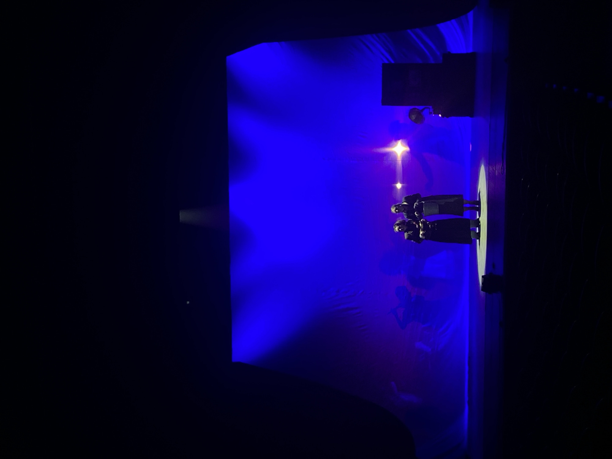 The image depicts a dimly lit stage, dominated by blue and purple lighting, with three people standing in the center, facing away from the camera. The people are dressed in dark clothing, and their features are not clearly visible. The stage's backdrop appears to be a cloth or fabric with undulating patterns. There is a stage light shining on the people. The location is potentially Rusanovo, Russia. The people are likely performing or preparing to perform on the stage. The environment is dark, with the stage lights providing the only illumination.