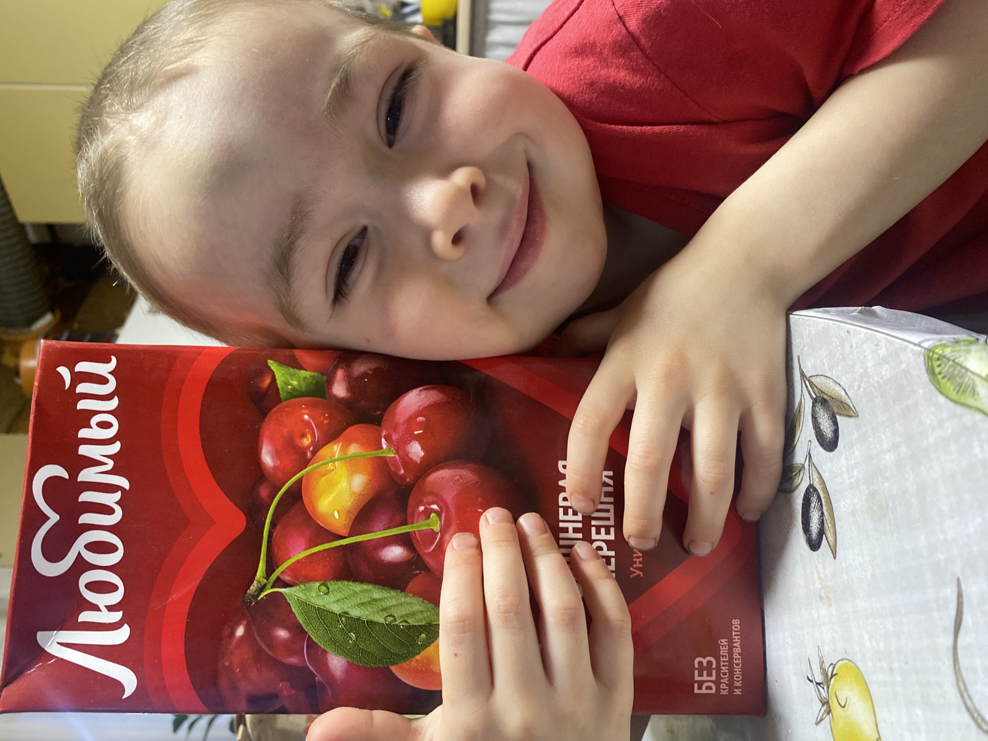 A child lies on a table in a room. The child is fair-skinned, with blond hair and wearing a red shirt. The child’s head is resting on a carton of cherry juice, and their hands are on the carton as well. The juice carton is red and features an image of cherries. The table has a patterned tablecloth. The image was taken indoors in a bright environment. The carton of juice indicates it is from Russia. The location, Ольховка, Russia, is based on the image context.