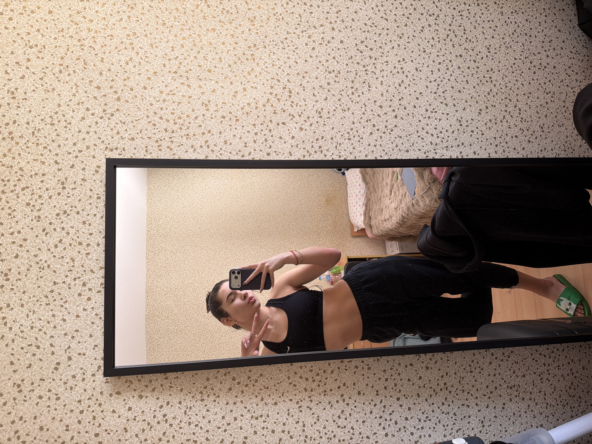 A full-length mirror selfie, presented rotated 90 degrees clockwise, captures a young woman posing playfully in an indoor setting. The woman, who appears to be in her late teens or early twenties, has dark hair tied back. She is dressed in a black sports bra, with a faint "Nike" logo visible on the front, and black sweatpants. She wears red bracelets on her right wrist and a delicate necklace.
She holds a smartphone with a dark case in her right hand, positioned to take the selfie, while her left hand forms a peace sign. Her expression is a pout or a blown kiss directed towards the camera.
The mirror's reflection shows a room with plain light-colored walls and a wooden floor. A bed, covered with beige and possibly patterned bedding, is partially visible behind the woman. On her feet, she wears distinctive green slide sandals adorned with a white cartoon character design.
The mirror itself has a slim black frame and is mounted against a wall characterized by a light beige or cream textured wallpaper, featuring numerous small, irregular, darker speckles. Part of a cleaning device, such as a vacuum or mop handle, is visible at the bottom edge of the image, outside the mirror's frame. The scene conveys a casual and self-assured atmosphere, likely within a personal living space in Sofia, Bulgaria.