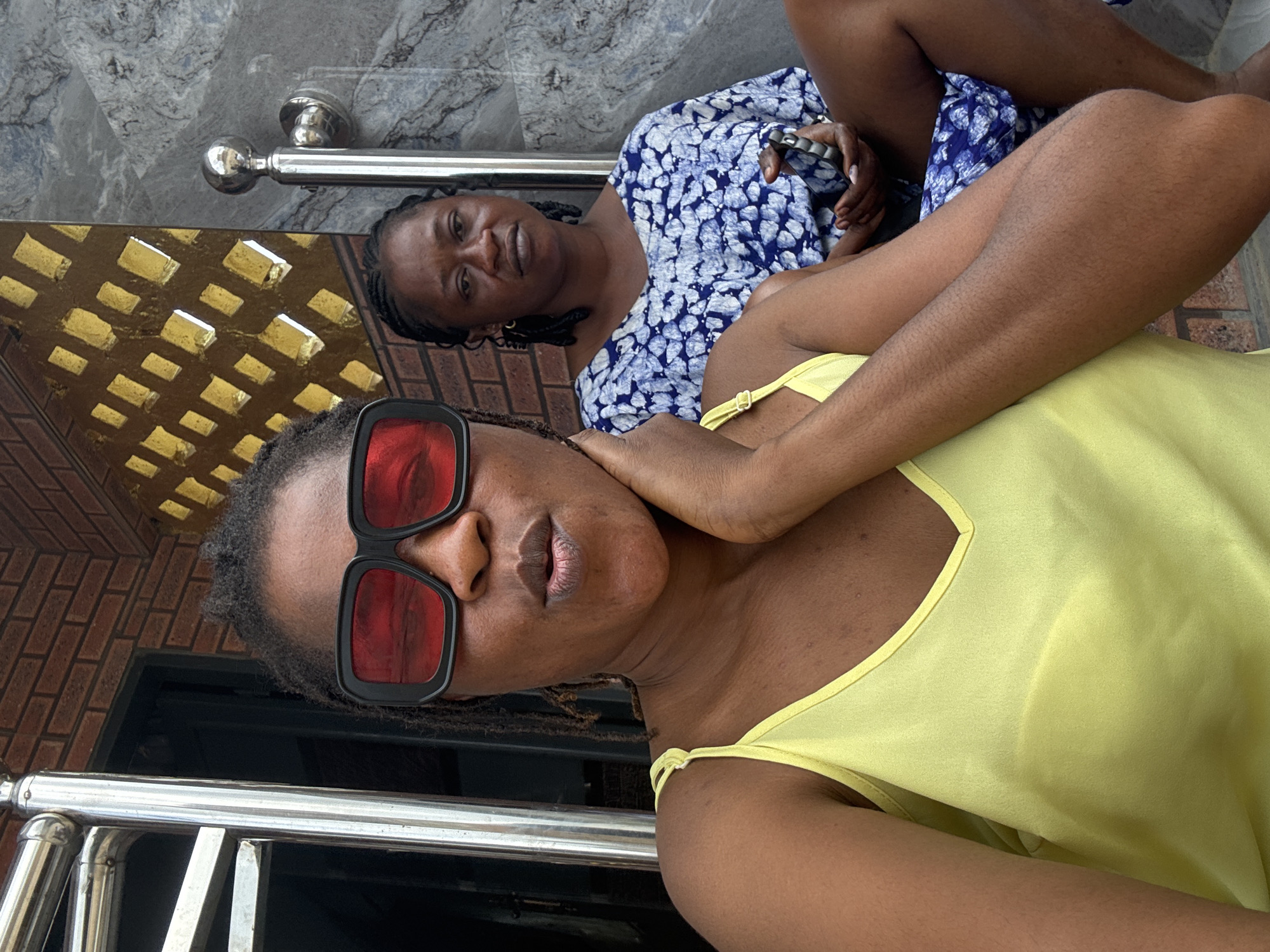 Two individuals are seated on a ledge outside a building with brick and marble facade. The person in the foreground, wearing a yellow top and large red-tinted sunglasses, is taking a selfie. Their left hand is resting on their neck. Behind them, a second person, wearing a blue and white patterned top, looks towards the camera holding a mobile phone. The environment suggests a residential setting.