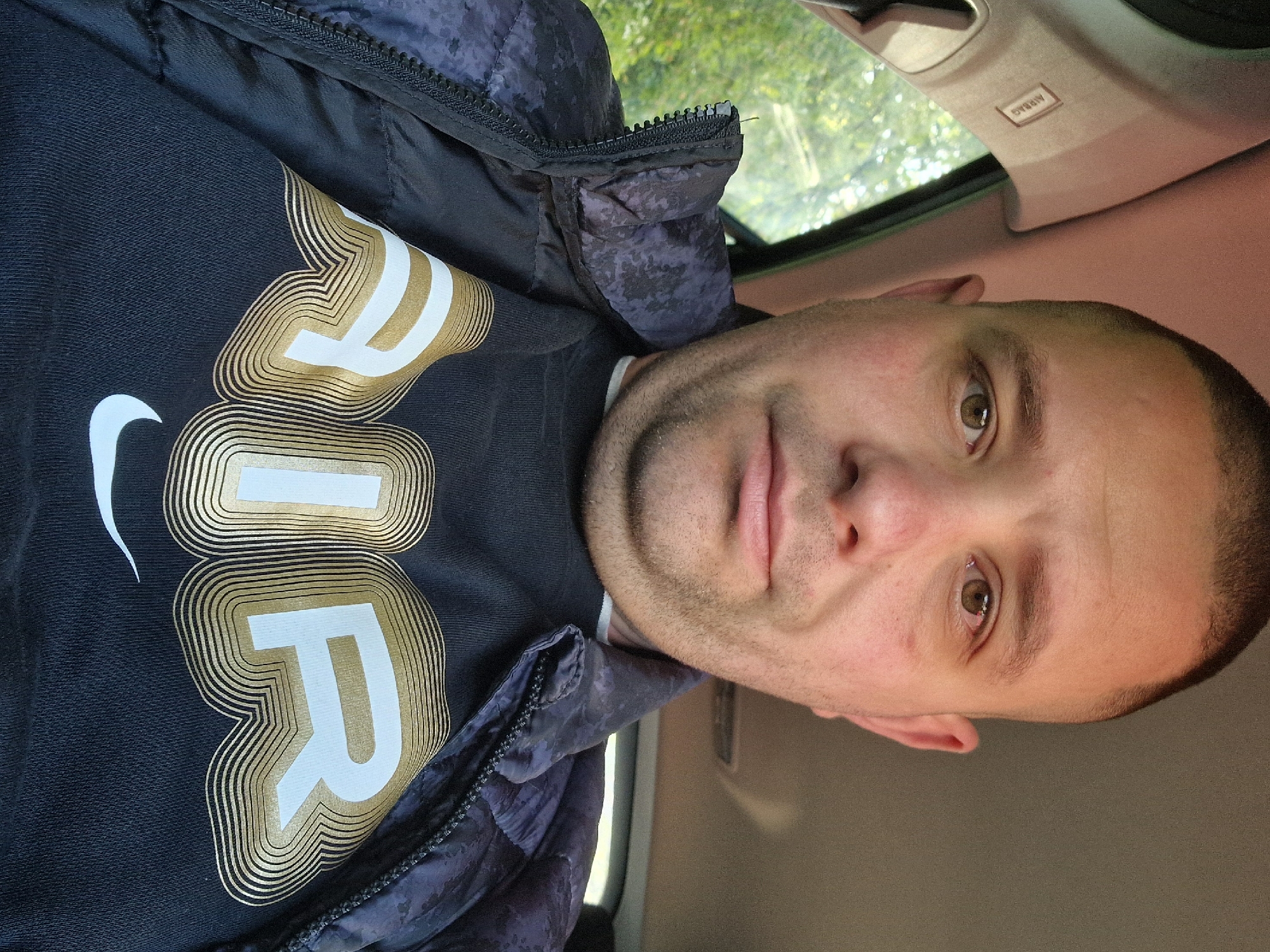 A medium shot captures a man inside a vehicle. He is wearing a black t-shirt with "AIR" written in white with gold accents, and a dark jacket. The interior of the car is visible, with an "AIRBAG" label on a panel above him. Outside the car window, a blurry view of foliage suggests an outdoor setting. There's no explicit evidence to pinpoint the location to Bearpark, United Kingdom in this image. The shot focuses mainly on the man and the interior of the vehicle.
