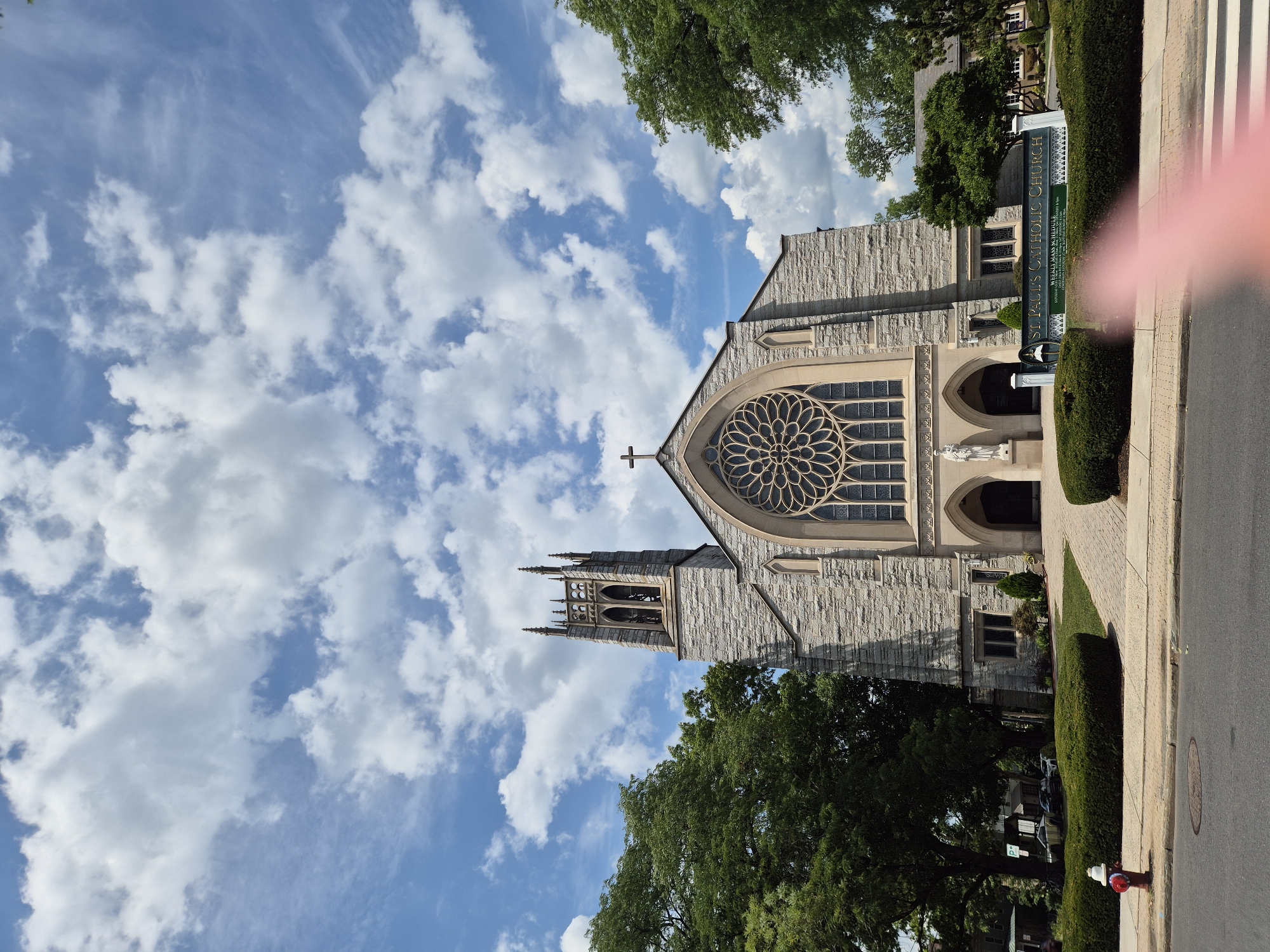 A wide-angle, low-angle, exterior shot shows the front of St. Paul's Catholic Church in Princeton, United States of America, under a bright blue sky with scattered white clouds. The church is constructed of light-colored stone and features a prominent bell tower on the left, a large rose window above the main entrance, and arched doorways. A statue stands to the left of the entrance. To the right of the entrance, a green sign with white lettering reads "ST. PAUL'S CATHOLIC CHURCH WEEKLY MASS SCHEDULE" followed by smaller text. The church is flanked by mature green trees and manicured hedges. In the foreground, a paved walkway leads to the church entrance. A red and white fire hydrant is visible on the left side of the sidewalk, next to a street. Part of a pink-toned object, possibly a finger, is partially obscuring the bottom right corner of the image. Cars are parked in the background on the left side of the church.