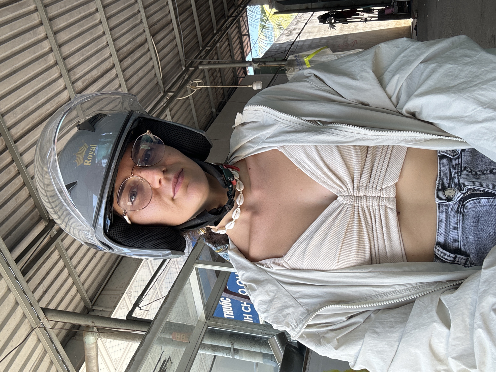 A young woman, wearing a gray helmet with a clear visor and a tan, ruched crop top, and a light-colored jacket, poses for a selfie under a corrugated metal roof structure. She has on glasses and a shell necklace. The background suggests an outdoor, urban setting with some construction or industrial elements, possibly in Hong Son, Vietnam, indicated by Vietnamese text on a sign. Sunlight filters through the structure.