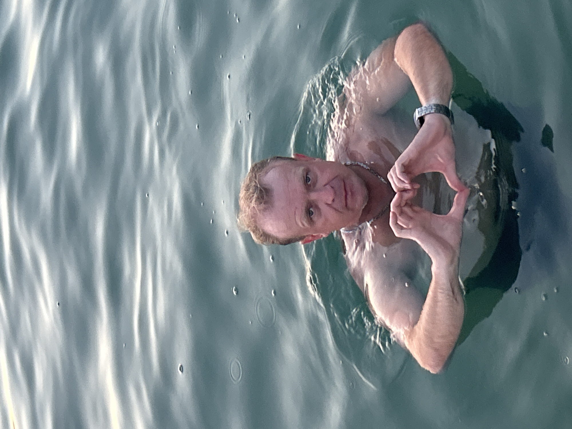 A man is in the water, forming a heart shape with his hands. He is looking at the camera with a neutral expression. The water is dark green and appears to be rippled. The man is wearing a watch on his left wrist. The scene is set in Katoro, Croatia.
