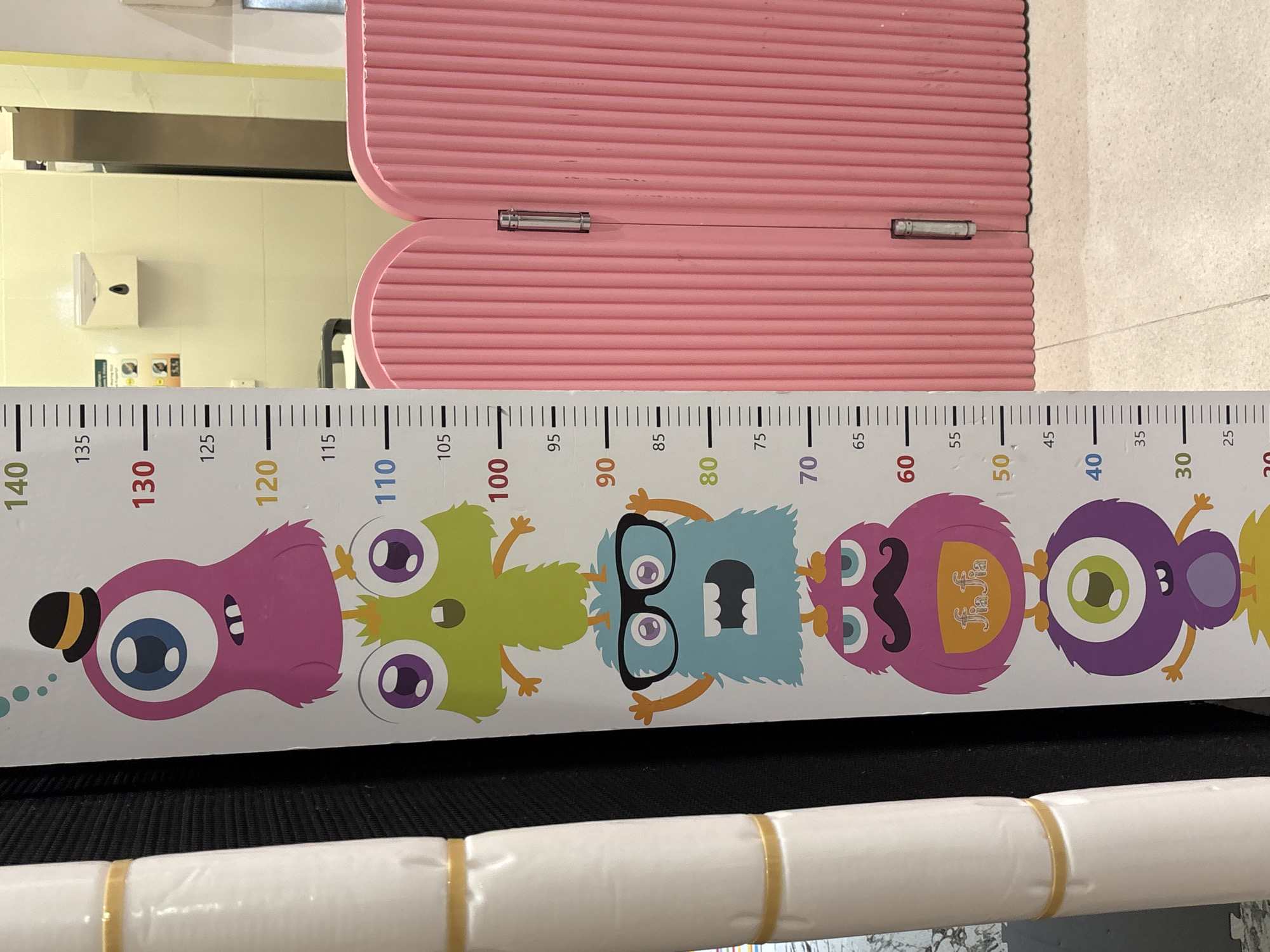 The image displays a portion of a height chart with cartoon monsters, set against a black background on the left and a pink plastic divider on the right. The height chart features markings from 20 to 140. Several cartoon monsters are stacked vertically on the chart, with the pink monster wearing a hat at the top, followed by a green monster, a blue monster wearing glasses, a pink monster with a mustache, and a purple monster with one eye at the bottom. The background on the right consists of a ribbed pink plastic divider that appears to be a child's play barrier, possibly in a public or commercial space. The floor beneath the divider is tiled. The overall scene suggests a child-friendly environment. There are no visible people in the image, and no specific landmarks identifiable to Dubai, United Arab Emirates.