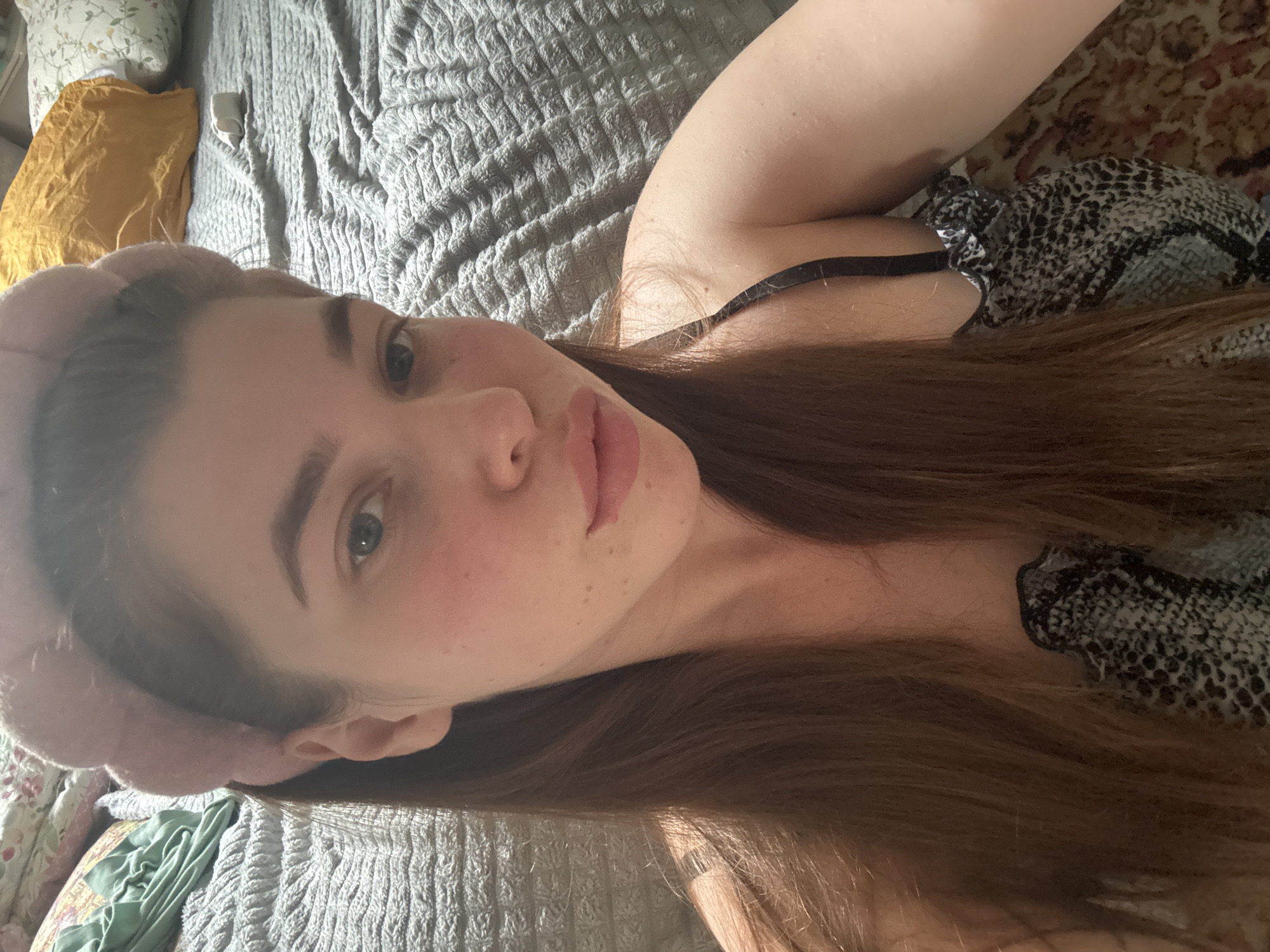 A woman is lying on a bed. She has long brown hair and is wearing a pink headband. She is wearing a black and grey top. The bed has a grey blanket and there is a yellow pillow and a pillow with floral design. The location is unknown, and cannot be determined from the image.