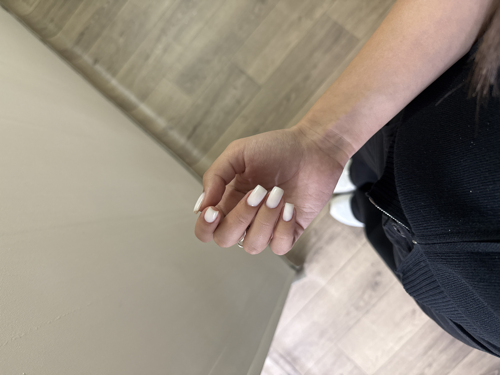 The image shows a person's left hand with square-shaped, white-colored nails. The hand is positioned against a backdrop of a light gray wall and light brown wood-grain flooring. The person is wearing black clothing and white sneakers. The image does not provide enough information to determine the location in Yerevan, Armenia.