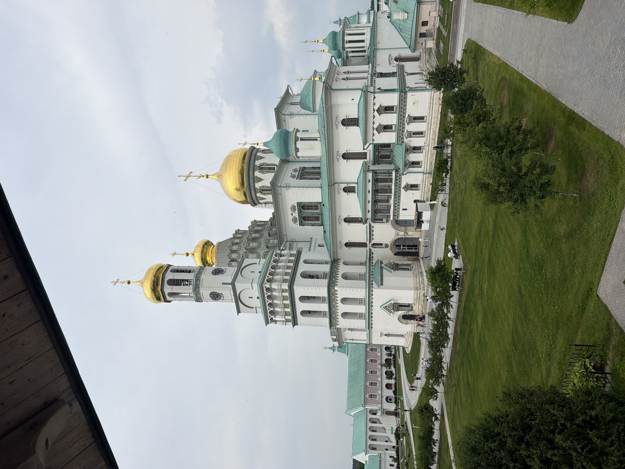 The image shows the Voskresensky Cathedral at the New Jerusalem Monastery in Istra, Russia. The cathedral is a large, white building with numerous golden domes and green accents. It is surrounded by a manicured lawn and paved walkways. Several people are walking and sitting on benches in the foreground. The sky is overcast. A wooden structure is visible in the upper left corner of the frame, obscuring part of the sky and the upper portion of the cathedral.