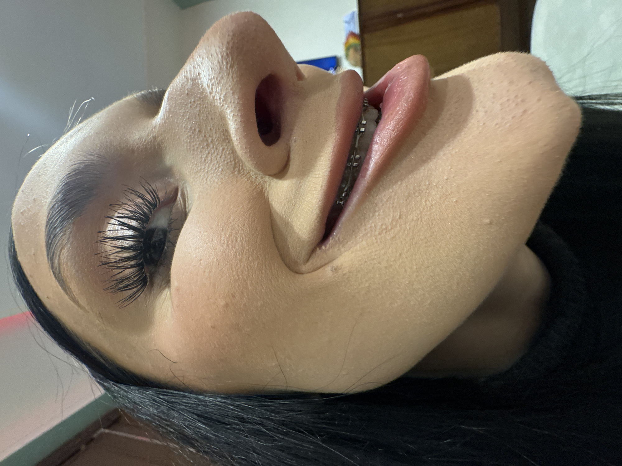 Here is a detailed description of the image:
**Scene:** A close-up, low-angle perspective of a person's face, likely taken indoors.
**People/Subject:**
* One person is visible, focusing on their face.
* They have fair skin, dark eyebrows, and noticeable false eyelashes.
* They are wearing braces on their teeth.
* Black hair is partially visible at the bottom of the frame.
**Objects:**
* Braces on the person's teeth.
* False eyelashes.
* Background objects are blurred, suggesting a room environment (furniture, possibly a wall).
**Actions/Interactions:**
* The person's head is tilted back, presenting the face in an unusual angle.
**Environment:**
* The environment is indoors.
* Location in the city of Unknown, Unknown.
* Background is blurred, not providing specific details.
* Lighting appears to be artificial and bright, possibly from an overhead source.