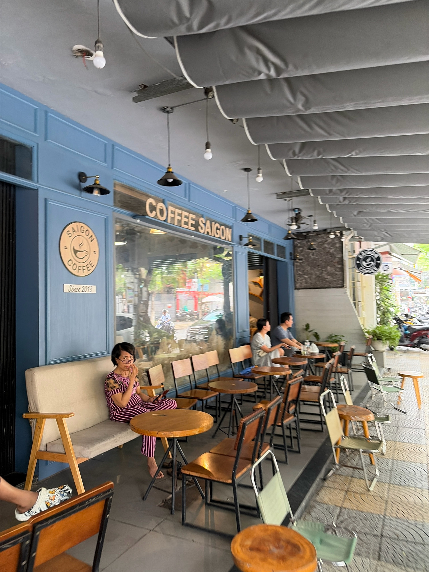 The image depicts the outdoor seating area of a coffee shop named "SAIGON COFFEE" located on a street in Đà Nẵng, Vietnam. The coffee shop features a blue facade with a large glass window. A circular logo with "SAIGON COFFEE" and "Since 2013" is visible on the left side of the building. Several tables and chairs are arranged on the tiled sidewalk.
In the foreground, a woman wearing striped pajamas is sitting on a light-colored sofa, holding a phone and looking at her hand. To her left, a white slide sandal with colorful butterflies is placed on the floor. Behind her, a woman and a man are seated at a table. Further back, other patrons are seen at tables, and a motorcycle is parked in the background. The scene is illuminated by natural daylight.