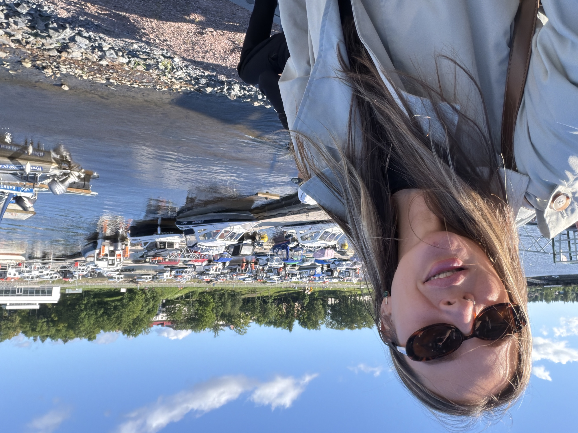 A young woman with long blonde hair and sunglasses is taking a selfie outdoors. She is wearing a light-colored trench coat over a black shirt. Behind her, a harbor is filled with numerous boats of various sizes docked in the water. The sky is blue with scattered clouds. The background shows a tree-lined embankment with buildings visible in the distance. The scene is the Sortavala waterfront in Sortavala, Russia.