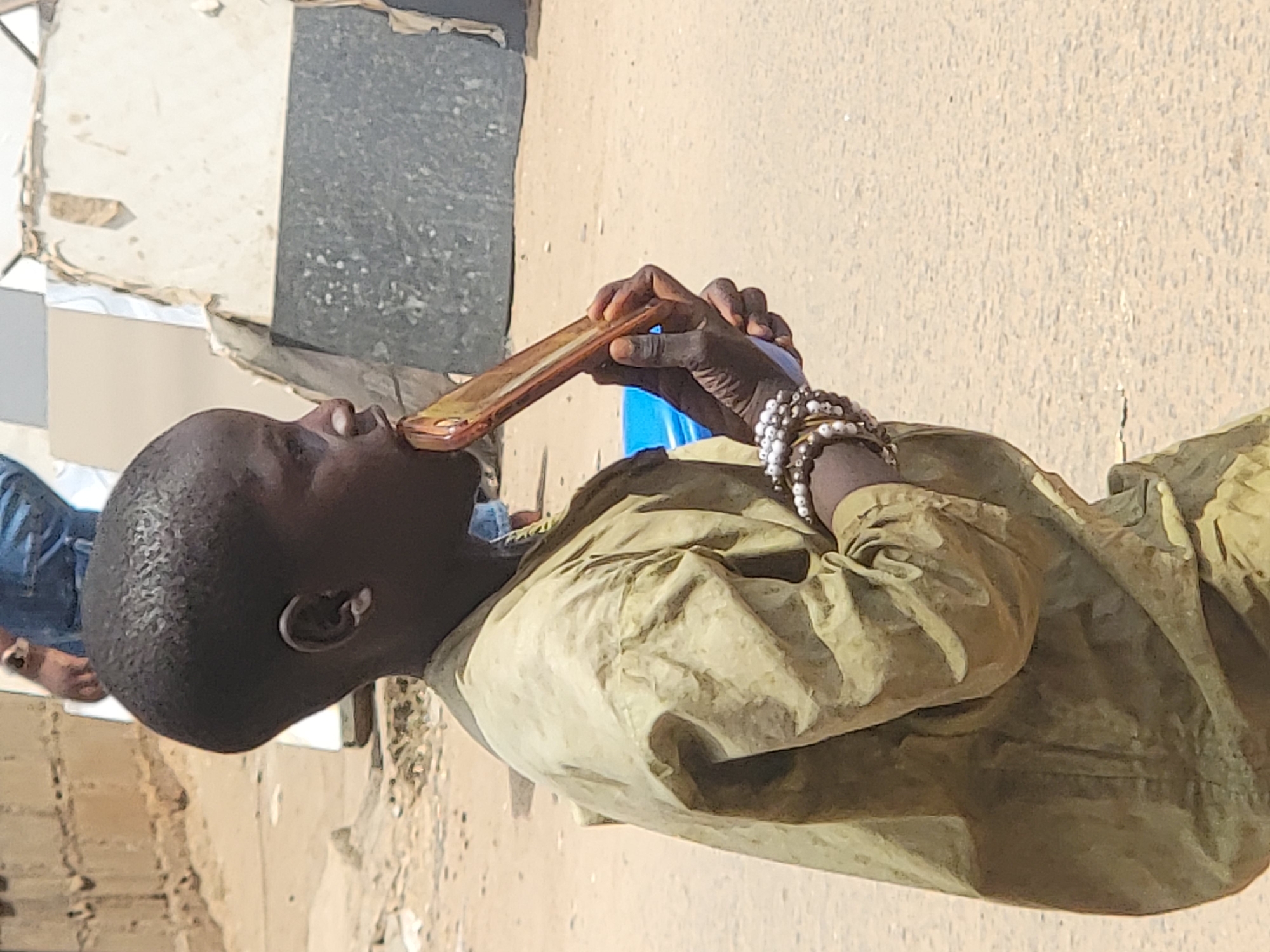 An African man is captured in an outdoor shot, seemingly within the city of Maiburiji, Nigeria. He holds a bronze-colored smartphone close to his mouth, possibly talking or recording. The man is wearing a khaki-colored collared shirt and multiple beaded bracelets. The background consists of a sandy ground, and partially visible buildings with a hint of blue sky in the distance. Another person's legs and feet are seen to the left of the man, implying a populated area.