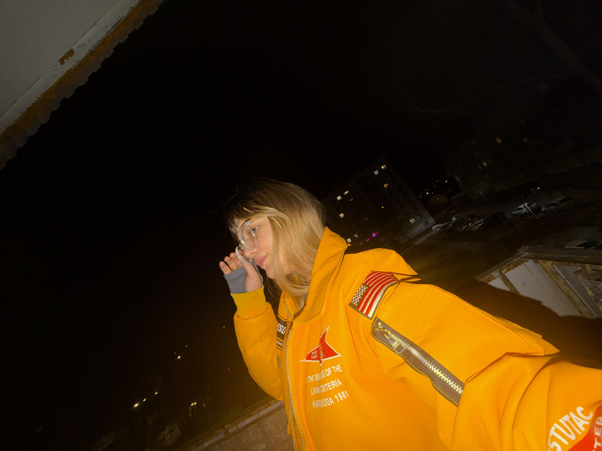 A woman stands outdoors at night, illuminated by an unseen light source. She wears glasses and a yellow jacket with American flag patches and text including "THE SHOES OF THE LIVING CRITERIA" and "HANSODA 1981."
In the background, a building with lit windows is visible. A city scene with cars and streetlights can be seen below the building. The location is Naberezhnye Chelny, Russia.