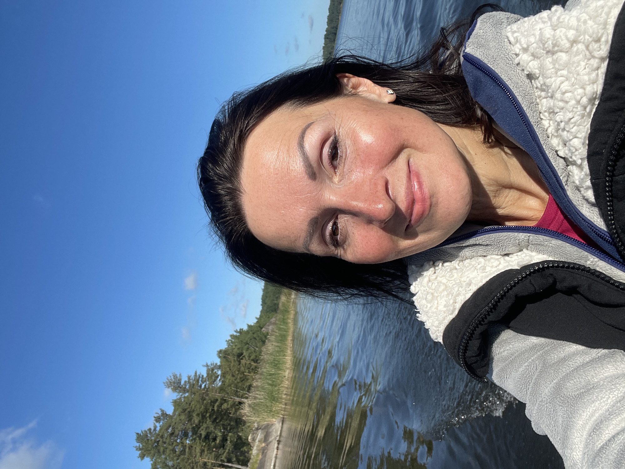 A woman is taking a selfie while on a boat. She has dark hair and is smiling. She is wearing a grey and white fuzzy jacket. The water is choppy, and there are trees and a blue sky in the background. There is no identifiable information to suggest the image is located in Berozovo, Russia.