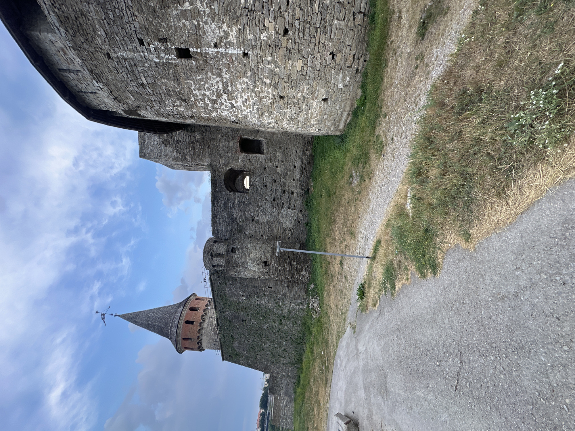 The image shows a section of the Kamianets-Podilskyi Castle in Kamianets-Podilskyi, Ukraine. The castle walls are constructed of rough-hewn stone, with some sections covered in moss and vegetation. A distinctive conical tower, topped with a weather vane, rises above the surrounding ramparts. The sky above is partly cloudy, with dramatic formations of white and grey clouds against a blue backdrop. In the foreground, a paved pathway, possibly a road or walkway, leads towards the castle. The area around the pathway is covered in dry grass and some scattered greenery. A single, light-colored metal pole stands near the pathway, suggesting the presence of lighting or signage. The overall scene depicts a historic fortification under a dynamic sky.