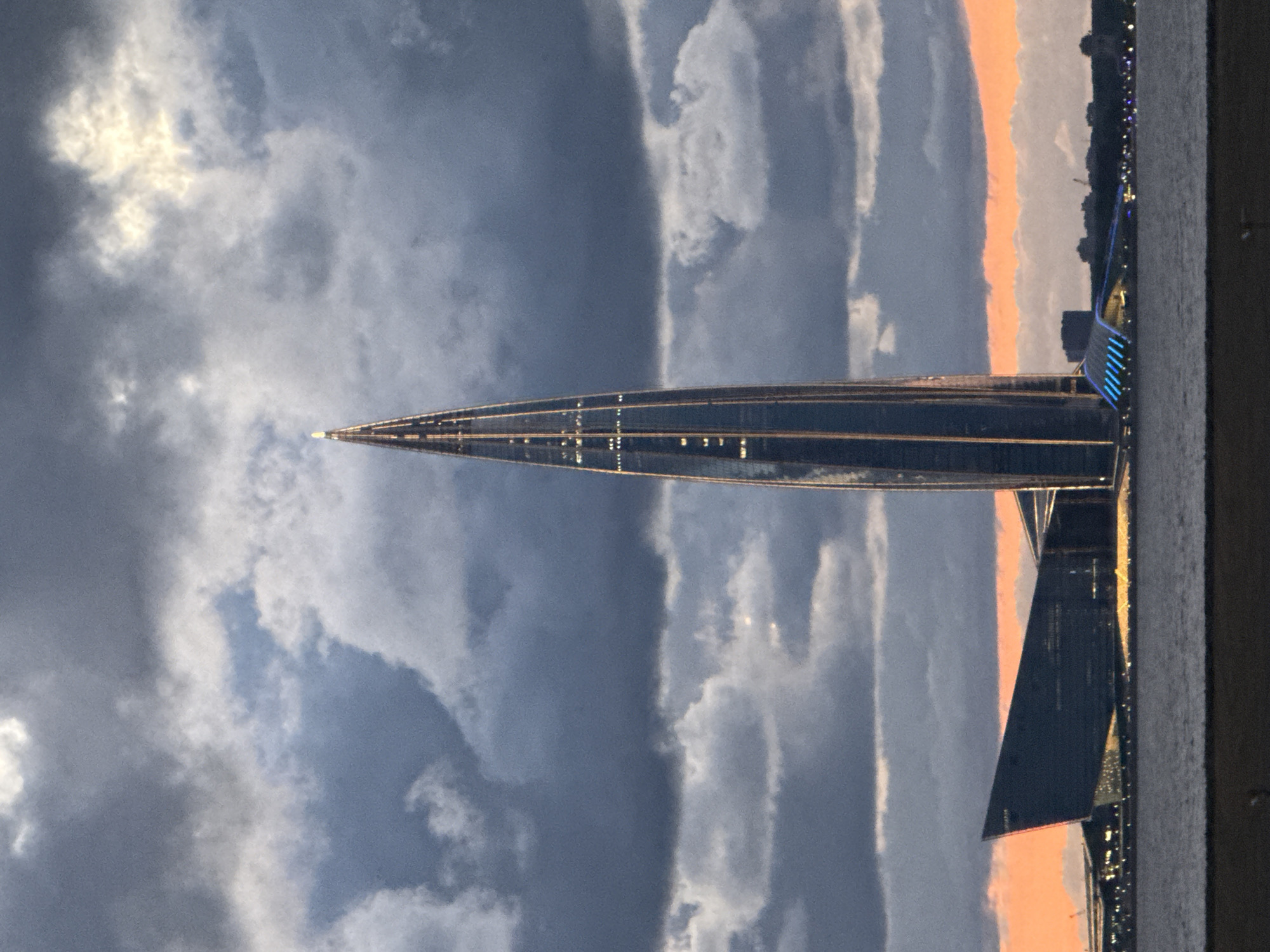 The image shows the Lakhta Center skyscraper in Saint Petersburg, Russia, on the shores of the Gulf of Finland. The skyscraper, with its distinctive spire, is illuminated by internal lights. In the foreground, the dark water of the gulf is visible, with a distant shoreline featuring other buildings and structures. The sky is dramatic and cloudy, with a band of orange light from the setting sun or sunrise visible near the horizon. The overall scene is one of architectural grandeur set against a moody natural backdrop.