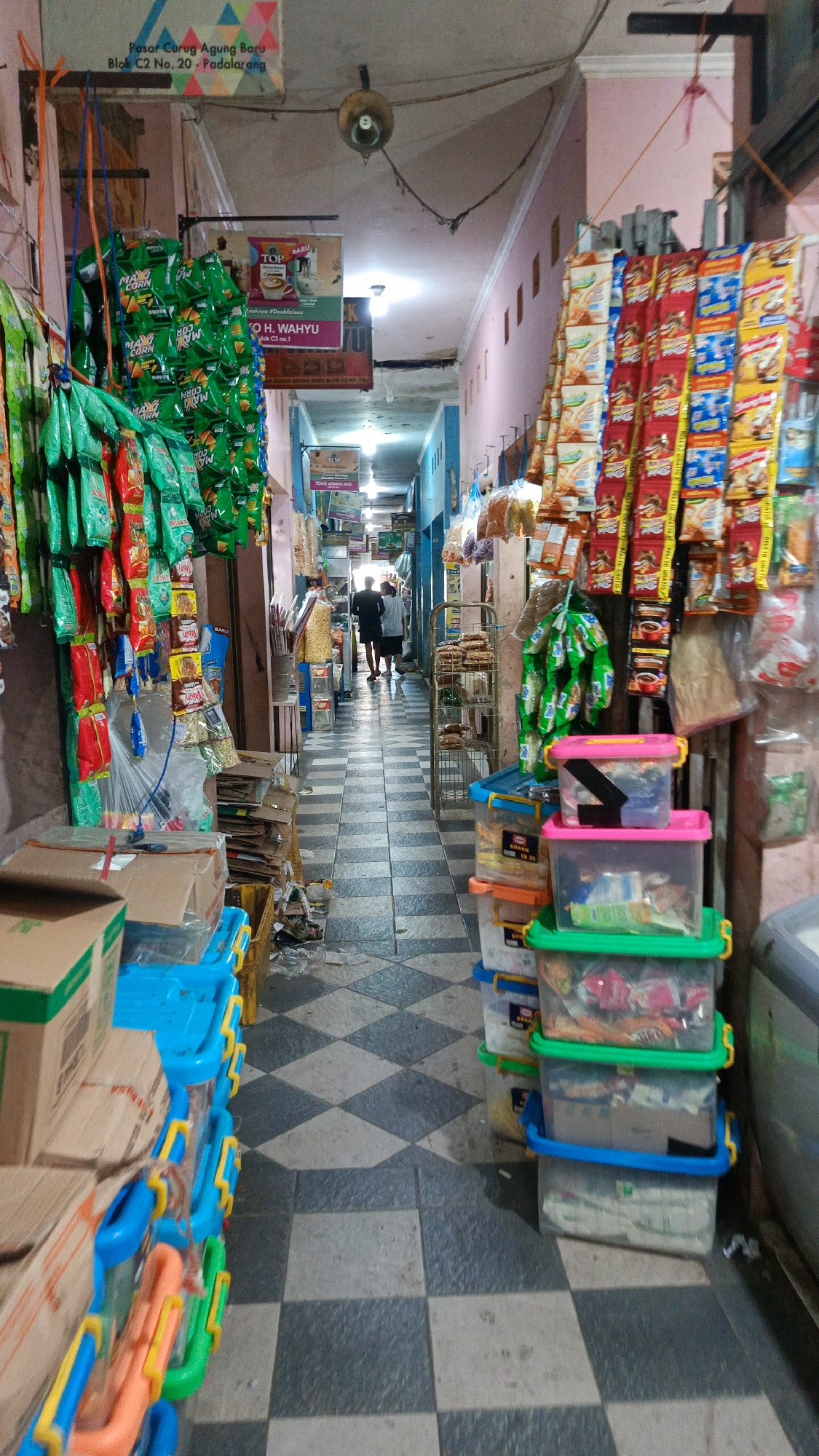 The image depicts a narrow indoor market aisle in Kertajaya, Indonesia. The aisle is lined with stalls selling various goods, including packaged snacks, drinks, and household items. The floor is tiled with a black and white checkered pattern. On the left, colorful bags of snacks hang from the ceiling, and stacks of boxes are piled near the wall. On the right, more packaged goods are displayed, along with clear plastic storage containers filled with items. Two individuals, appearing to be customers or vendors, are seen walking away from the camera towards the end of the aisle. The lighting suggests an indoor setting, possibly a covered market or a row of shops. Signs in Indonesian are visible, indicating the market's location and potentially the names of businesses. The overall scene conveys a busy, commercial atmosphere typical of local Indonesian markets.