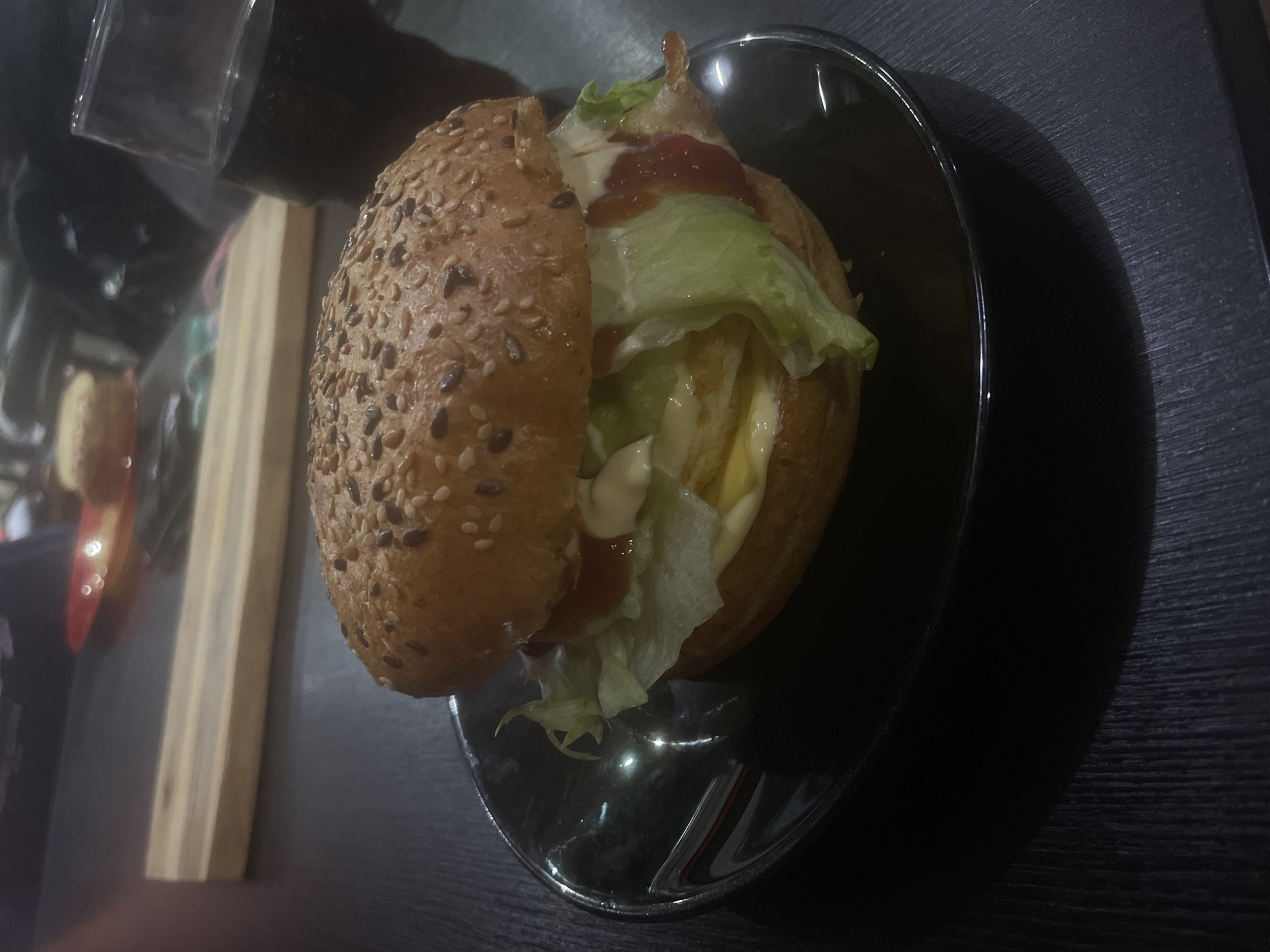 A sesame seed bun burger sits on a dark, glossy dish on a dark surface. The burger contains lettuce, mayonnaise, and ketchup. A glass and a wooden object are partially visible near the burger. The location is likely Burgas, Bulgaria.