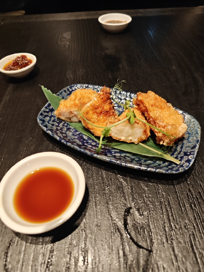 Four pieces of fried food are arranged on a blue patterned plate with a green leaf underneath. Garnishes are placed on top of the food. Small bowls of dipping sauce surround the plate. A shallow dish contains a red-brown liquid. A dark wooden table serves as the background.