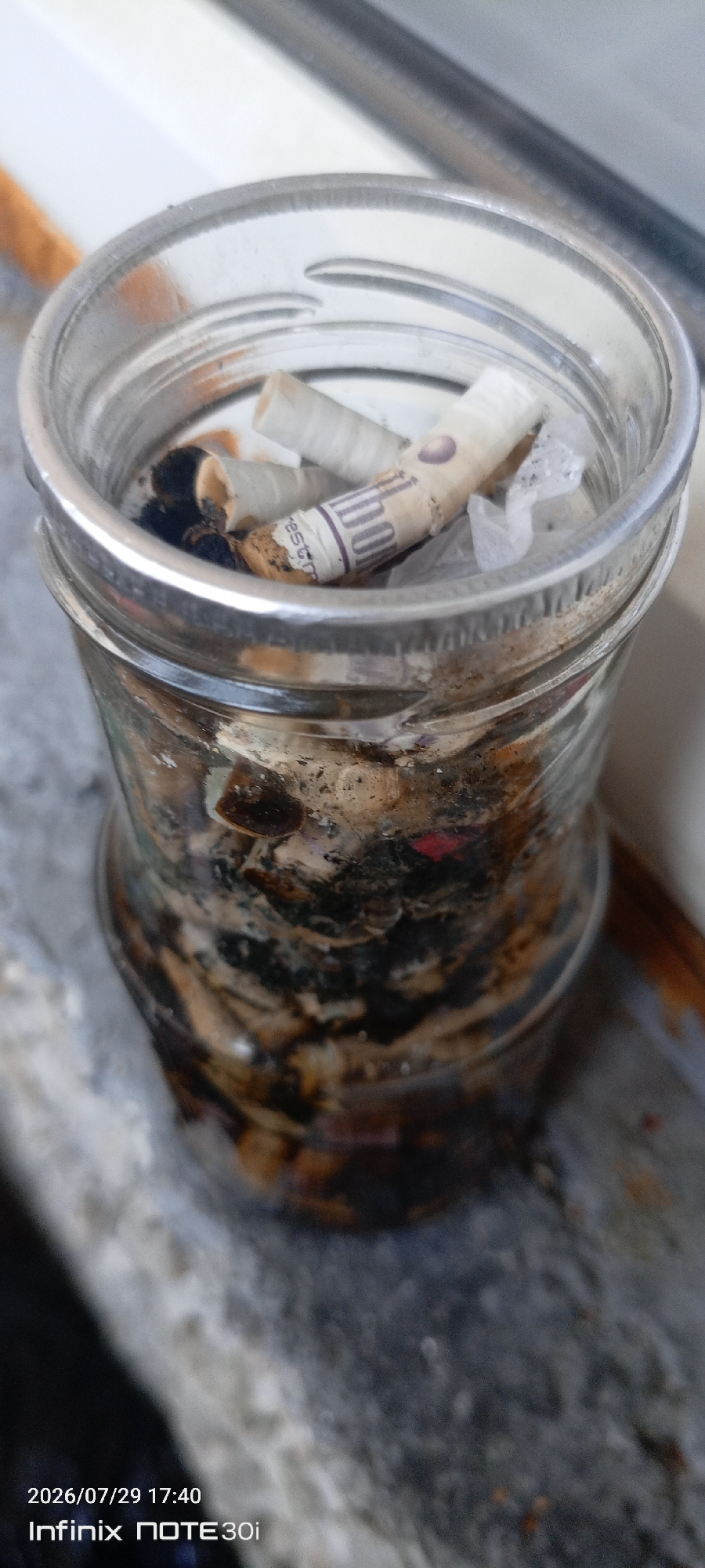 The image shows a close-up, eye-level view of a glass jar filled with cigarette butts. The jar appears to be made of clear glass with a metallic rim. The cigarette butts are numerous and appear to be accumulated, with ash and remnants of burnt tobacco visible. Some of the butts have white paper filters, while others show brown tobacco. In the top of the jar, there are a few cigarette butts that are more clearly visible, one of which has purple writing on it. The jar is placed on a ledge or windowsill, with the background showing a blurred, outdoor environment. The lighting suggests it is daytime. Visible text in the bottom left corner indicates the date "2026/07/29 17:40" and the device model "Infinix NOTE 30i". The setting appears to be a domestic or outdoor space, possibly a balcony or window area. No people are visible in the image. The overall impression is one of accumulated waste.