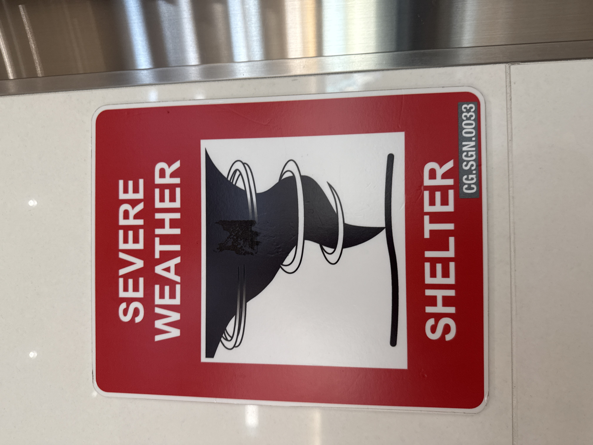 A "Severe Weather Shelter" sign is affixed to a white tiled surface, presumably on the floor of an indoor space. The sign is rectangular with rounded corners and features a white tornado graphic on a red background, with the phrase "SEVERE WEATHER" displayed vertically on the left side of the sign and the word "SHELTER" vertically on the right side, also in white lettering. "CG.SGN.0033" is printed in dark gray along the right edge of the sign. The image appears to have been taken indoors, as evidenced by the indoor lighting and tiled floor. The presence of the sign suggests that this is an area designated for shelter during severe weather events. Although the precise location cannot be determined from the image alone, the sign's potential existence within Fort Lauderdale, USA is noted in the prompt. No people are visible in the image, and no specific actions or interactions are taking place.